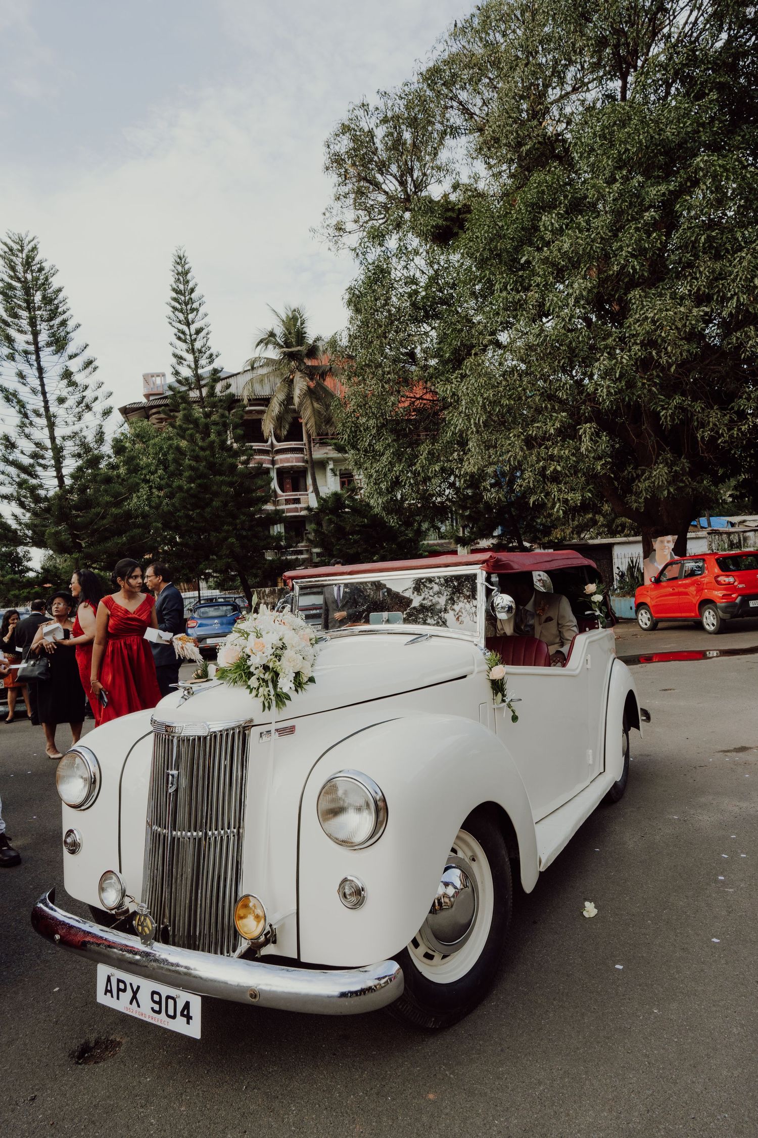 Vintage white convertible car decorated with flowers parked on a street while guests in colorful dresses gather beside tall green trees.