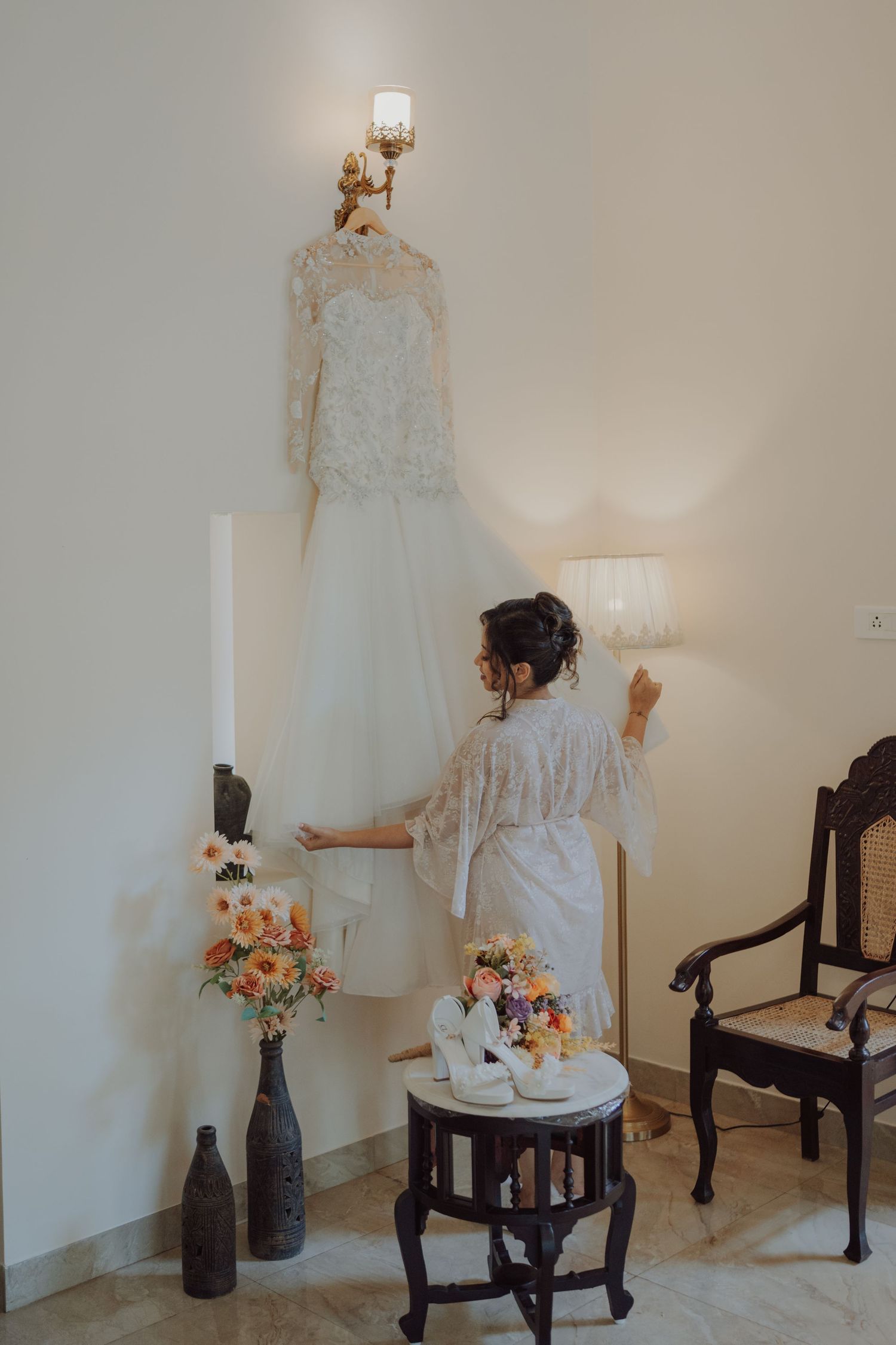 A bride in a lace robe touches her white wedding gown hanging on the wall beside a floral arrangement and shoes.