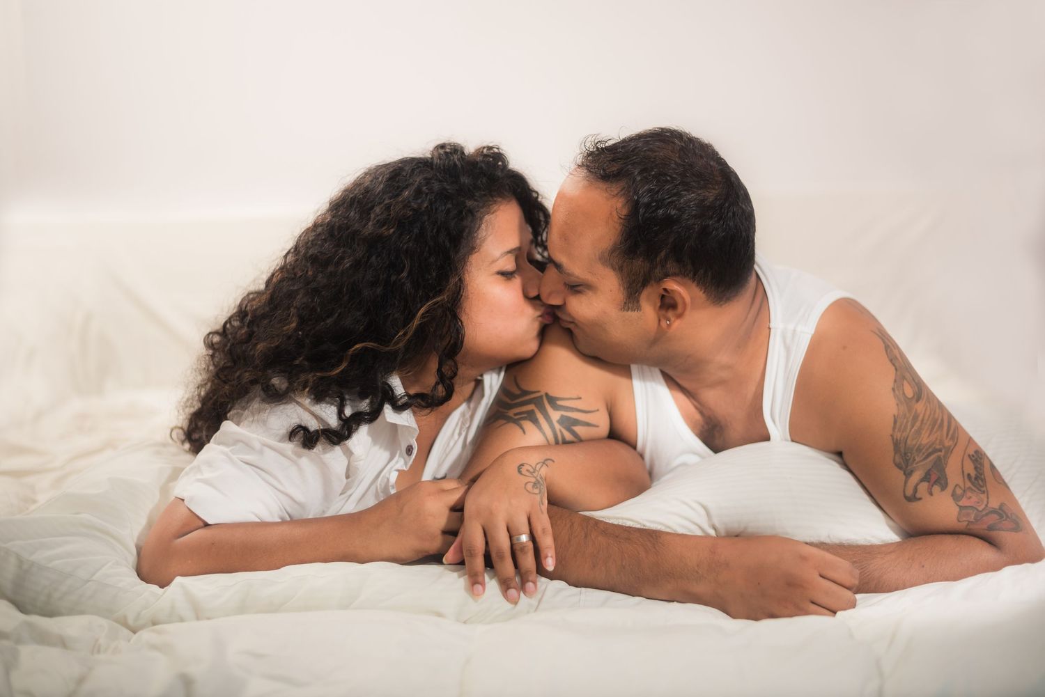 A couple shares an intimate moment while lying together on white bedding.