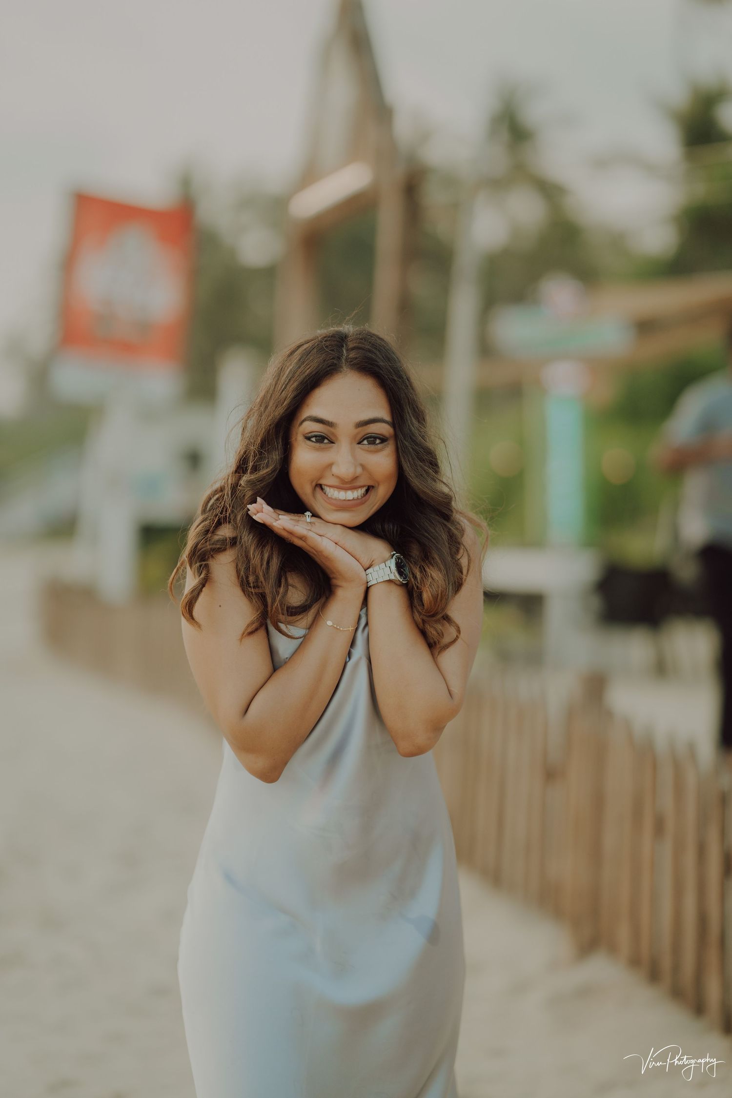 A person in a light gray dress poses outdoors with hands near face against a blurred tropical background.