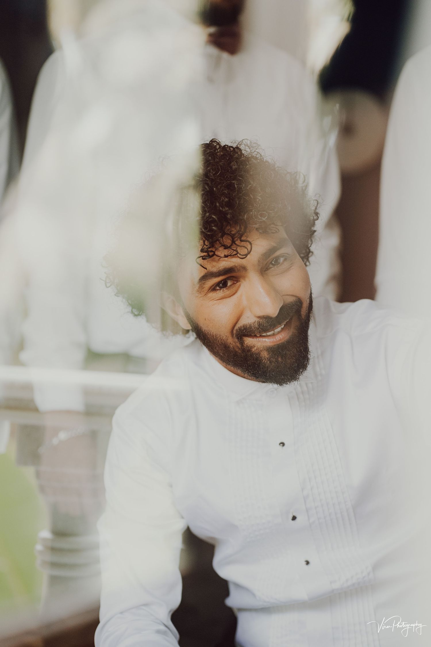 A smiling individual in a white button-up shirt poses for a soft-focus portrait with natural lighting.