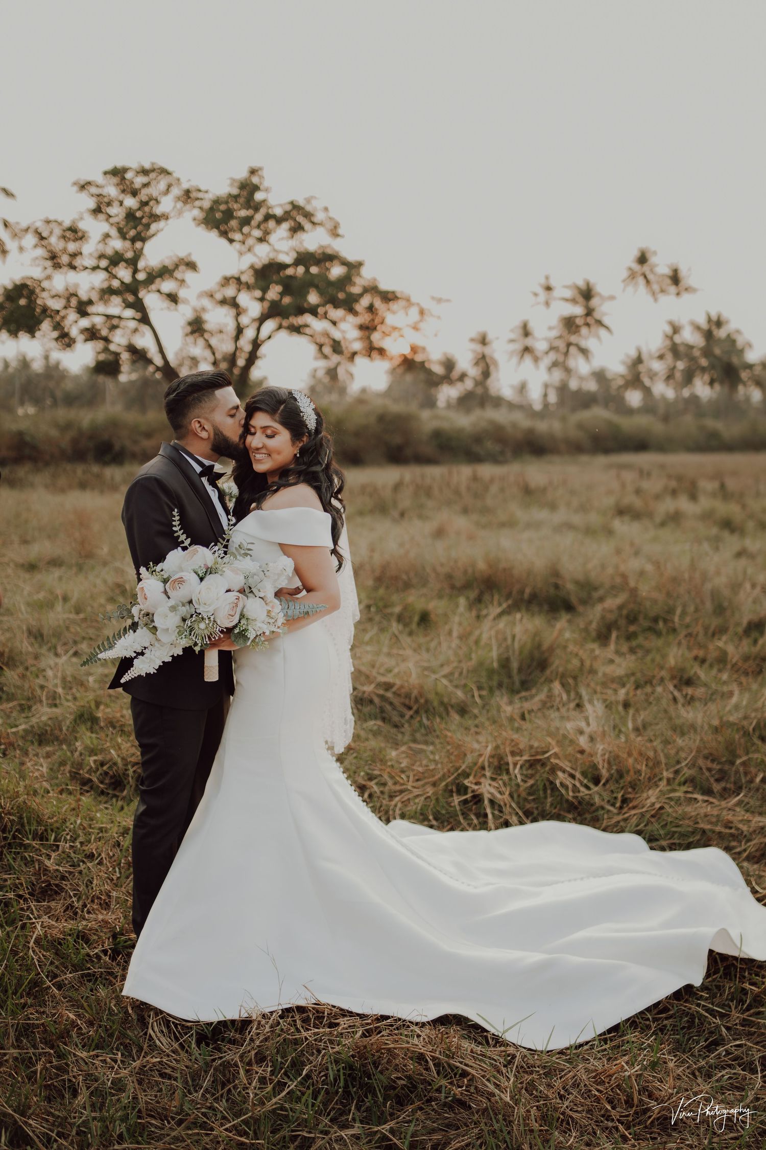 A couple shares a romantic moment in their wedding attire at sunset in a rustic field with flowing white gown and bouquet.