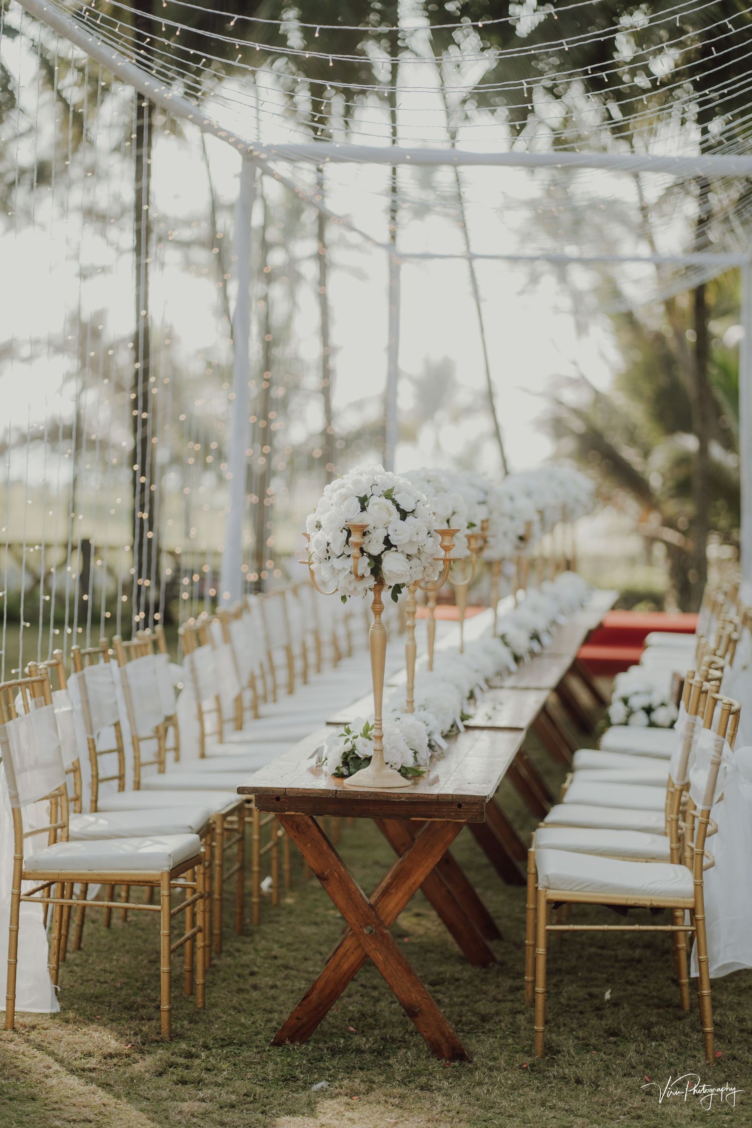 An elegant outdoor wedding reception setup with rustic wooden tables, bamboo chairs and hanging string lights under a tent canopy.