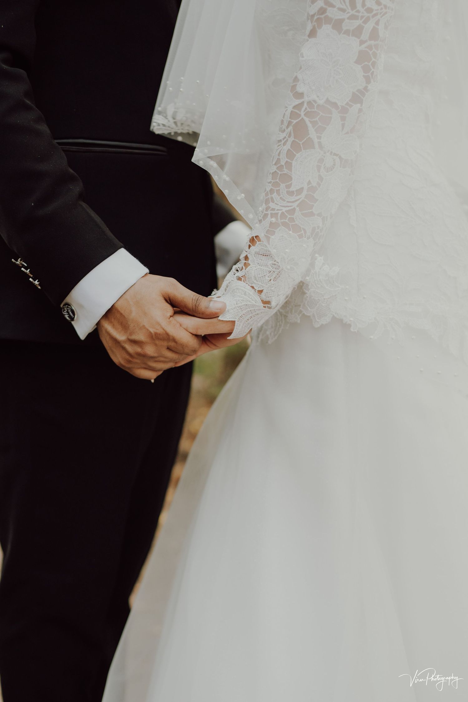 Close-up shot of couple holding hands at a wedding, showing black suit sleeve and white lace wedding dress details.