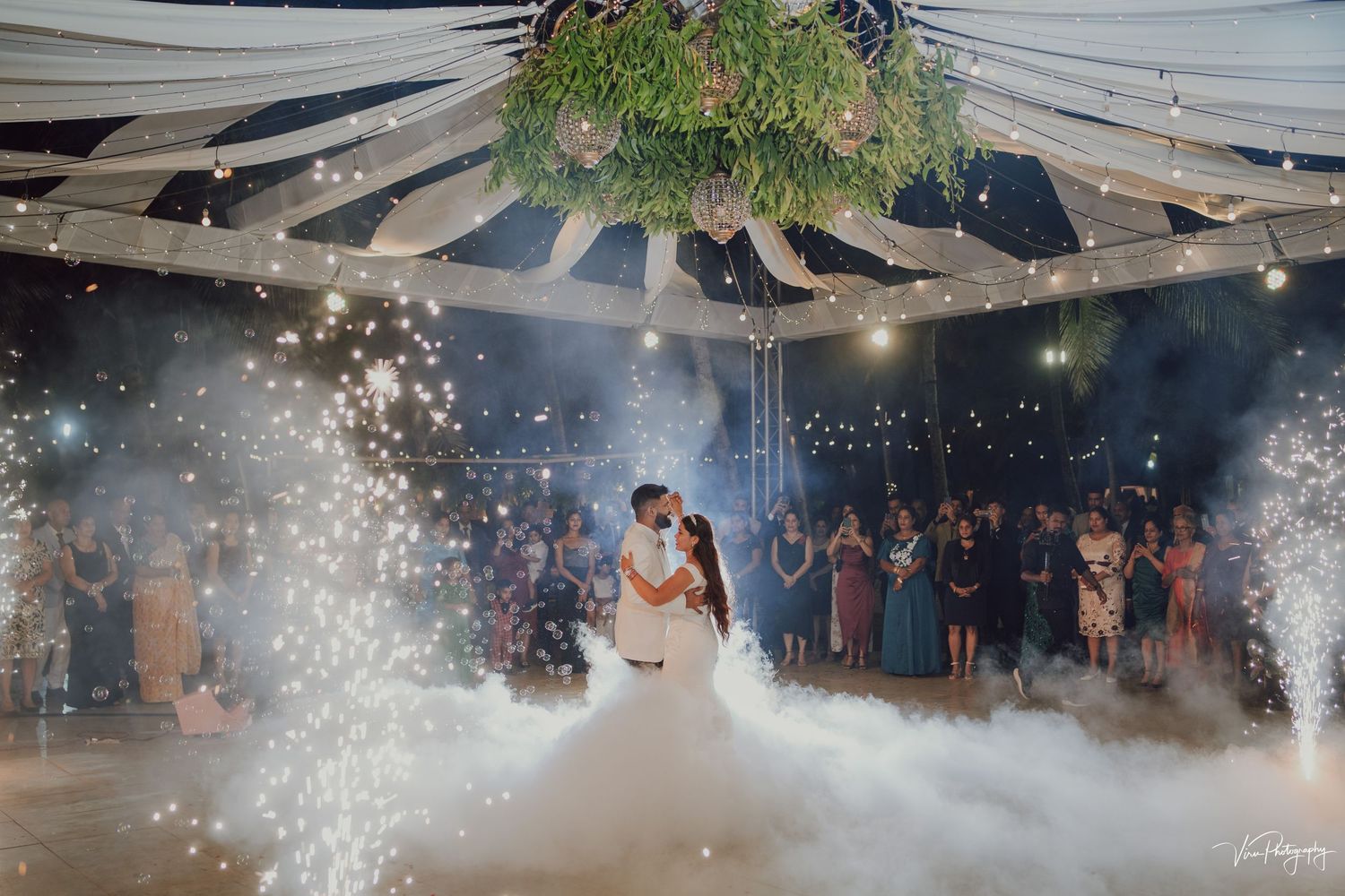 A couple shares their first dance at a wedding reception with sparklers and fog effects creating a magical atmosphere.