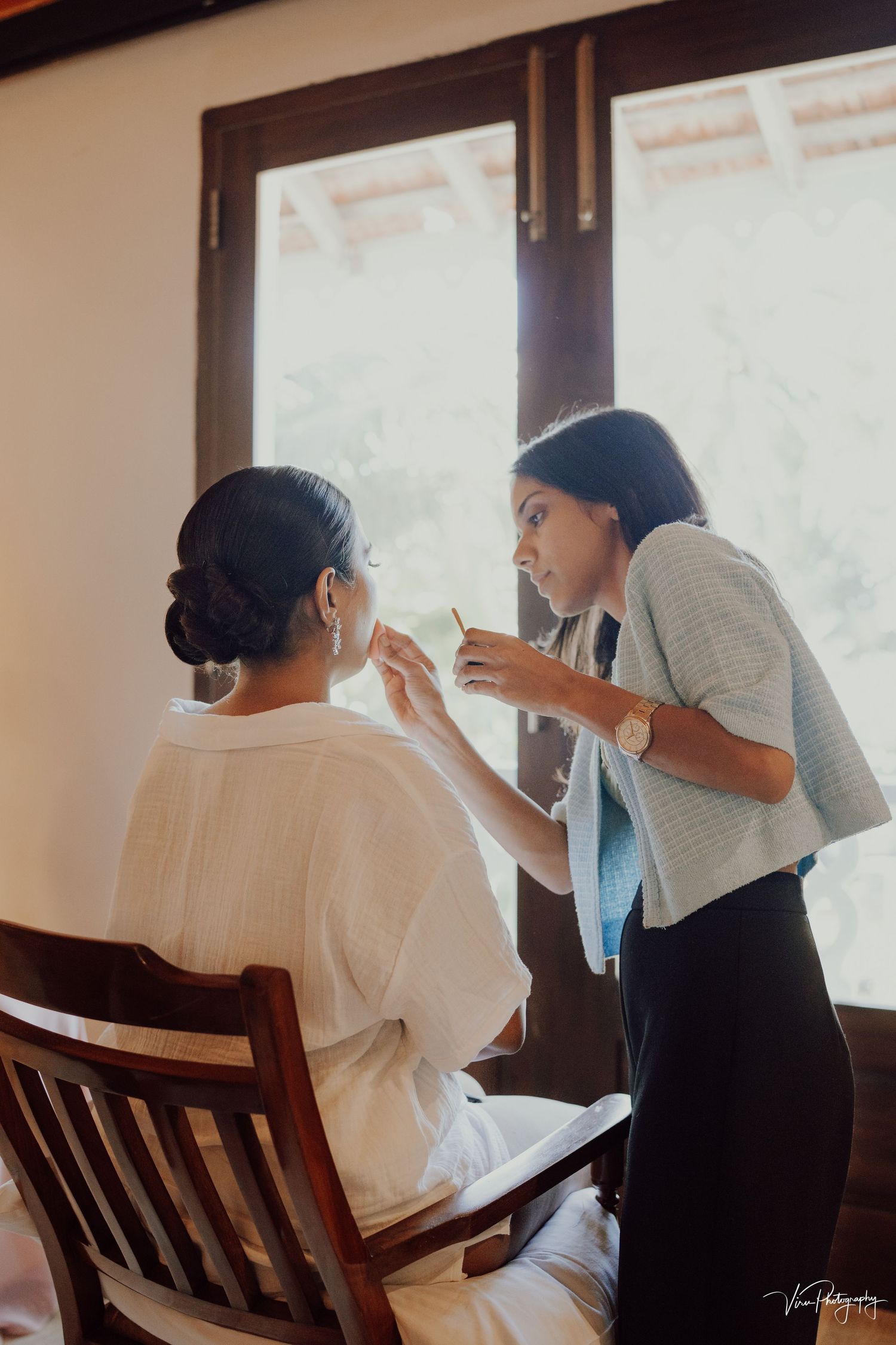 A makeup artist applying cosmetics to a client seated in a wooden chair by a window in warm lighting.