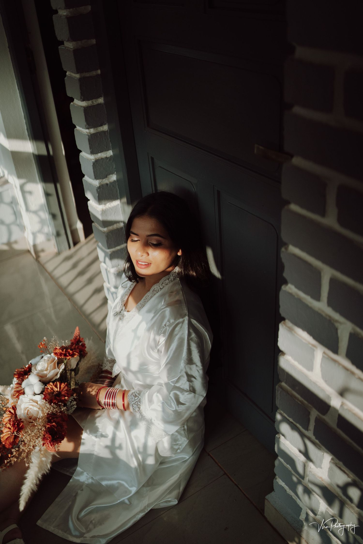 Moody portrait in white dress with autumn florals sitting by brick wall and door in dramatic lighting.