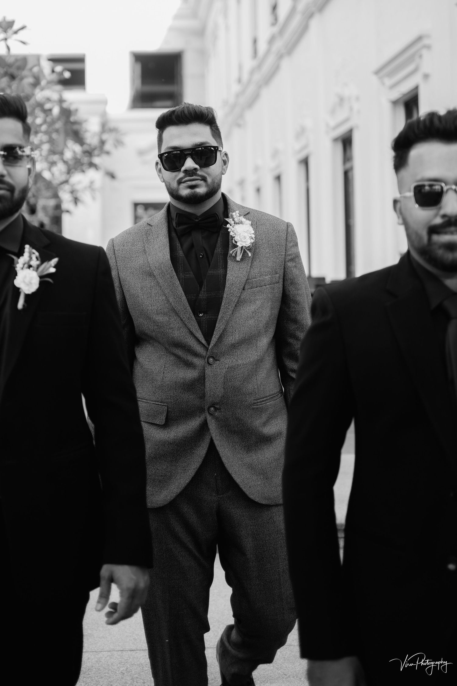 Black and white wedding photo of groomsmen wearing sunglasses and suits walking down a street together.