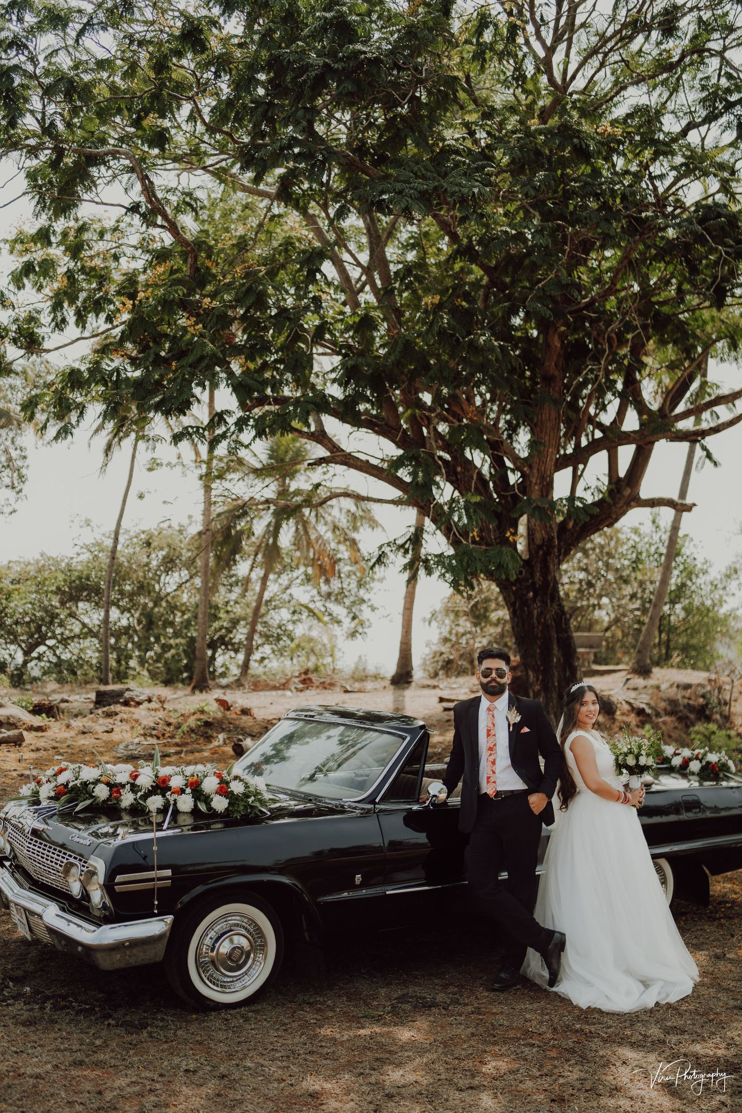 A vintage black Chevrolet Impala decorated for a wedding ceremony parked under a large tree in a rustic outdoor setting.