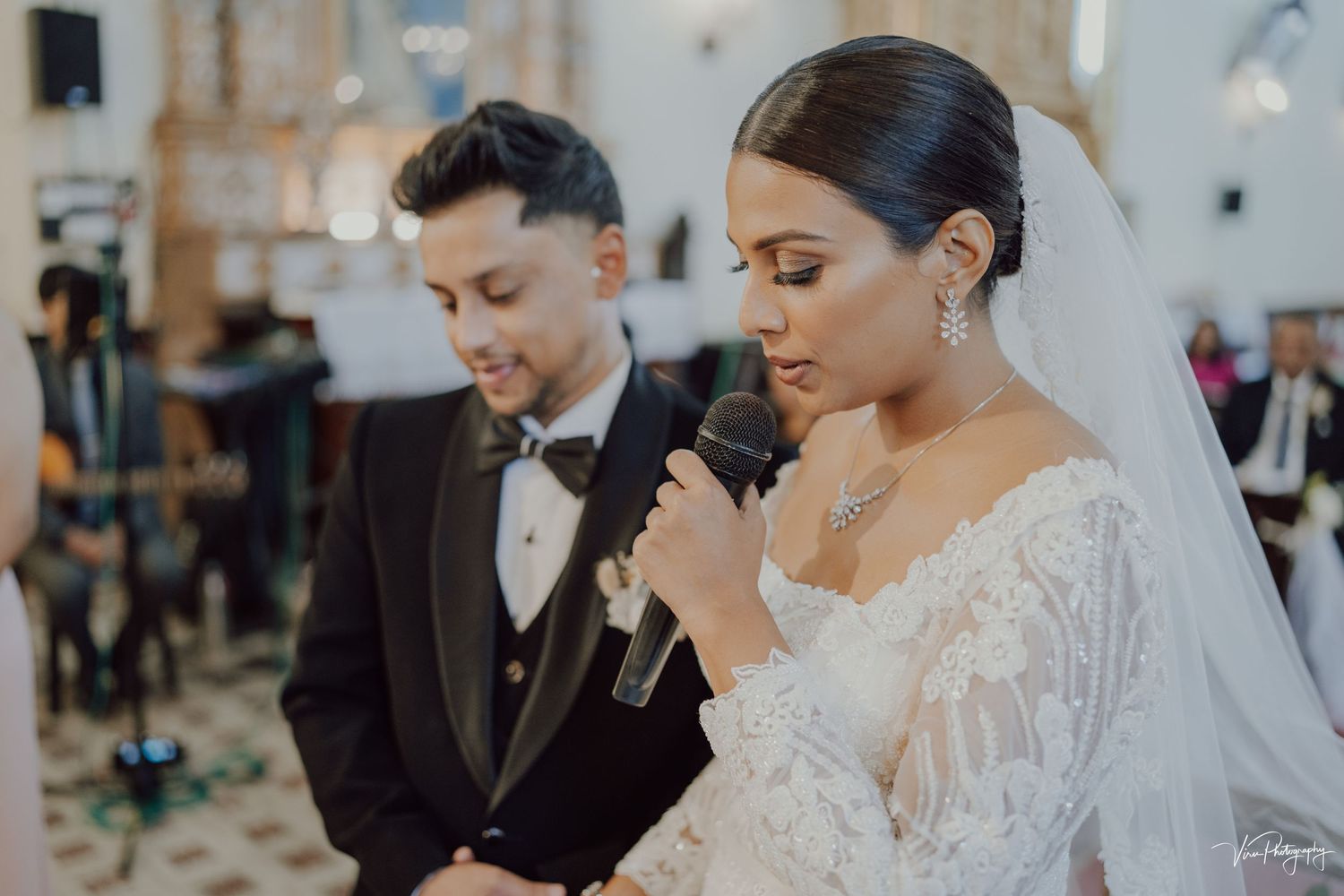 A bride in white lace dress and veil prays beside her groom in a black tuxedo during a church wedding ceremony.