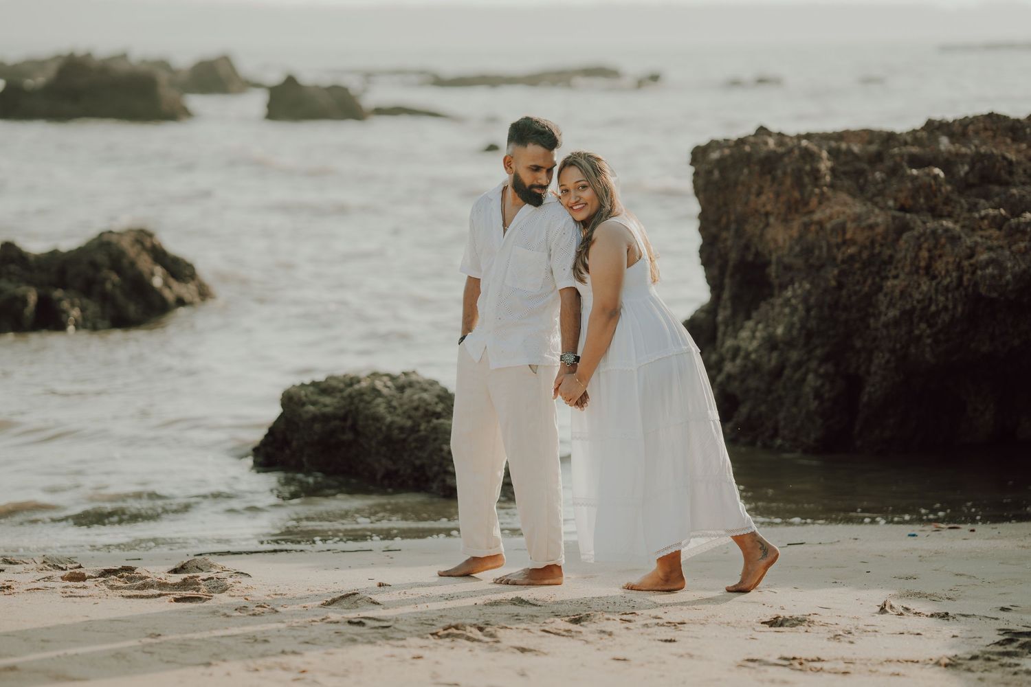 A couple in white attire shares intimate moments along a rocky beach during a romantic photoshoot at sunset.