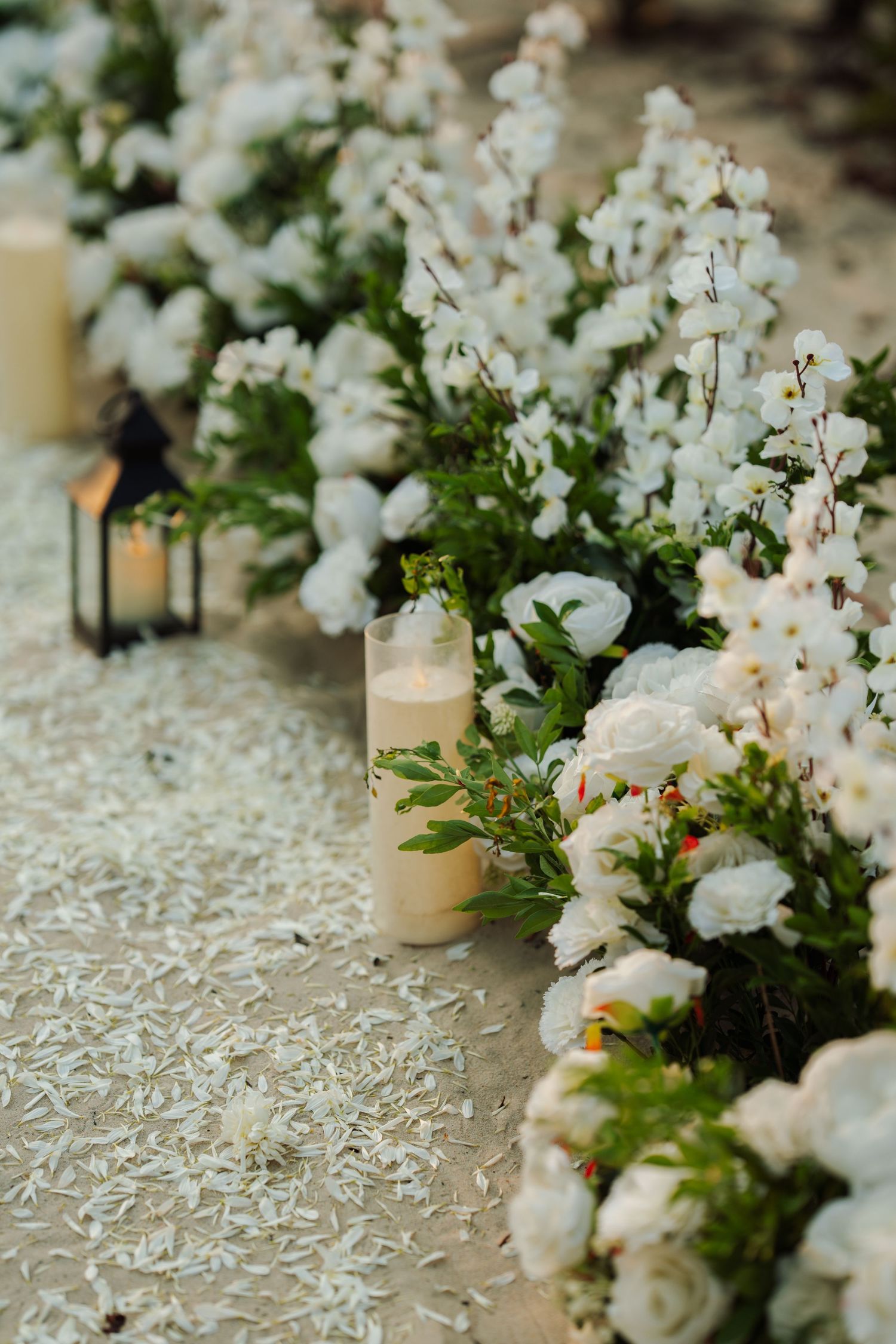 White floral wedding aisle with candles, green foliage, and scattered petals on sandy ground.