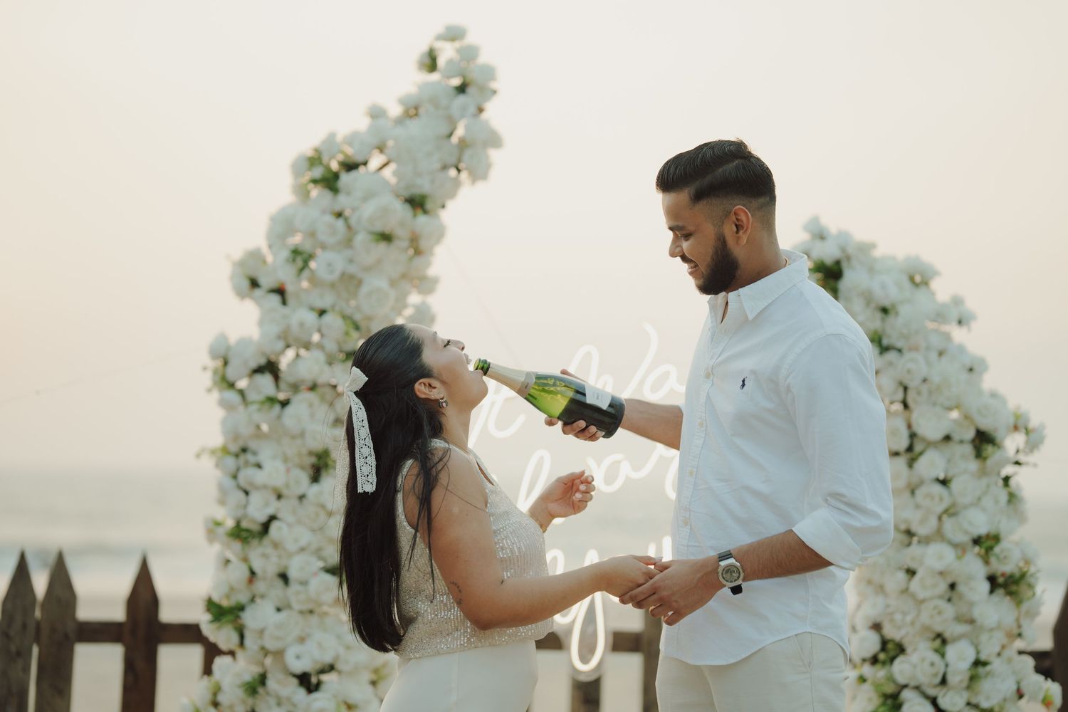 Couple in white attire celebrating with champagne under a floral arch on a beach at sunset.