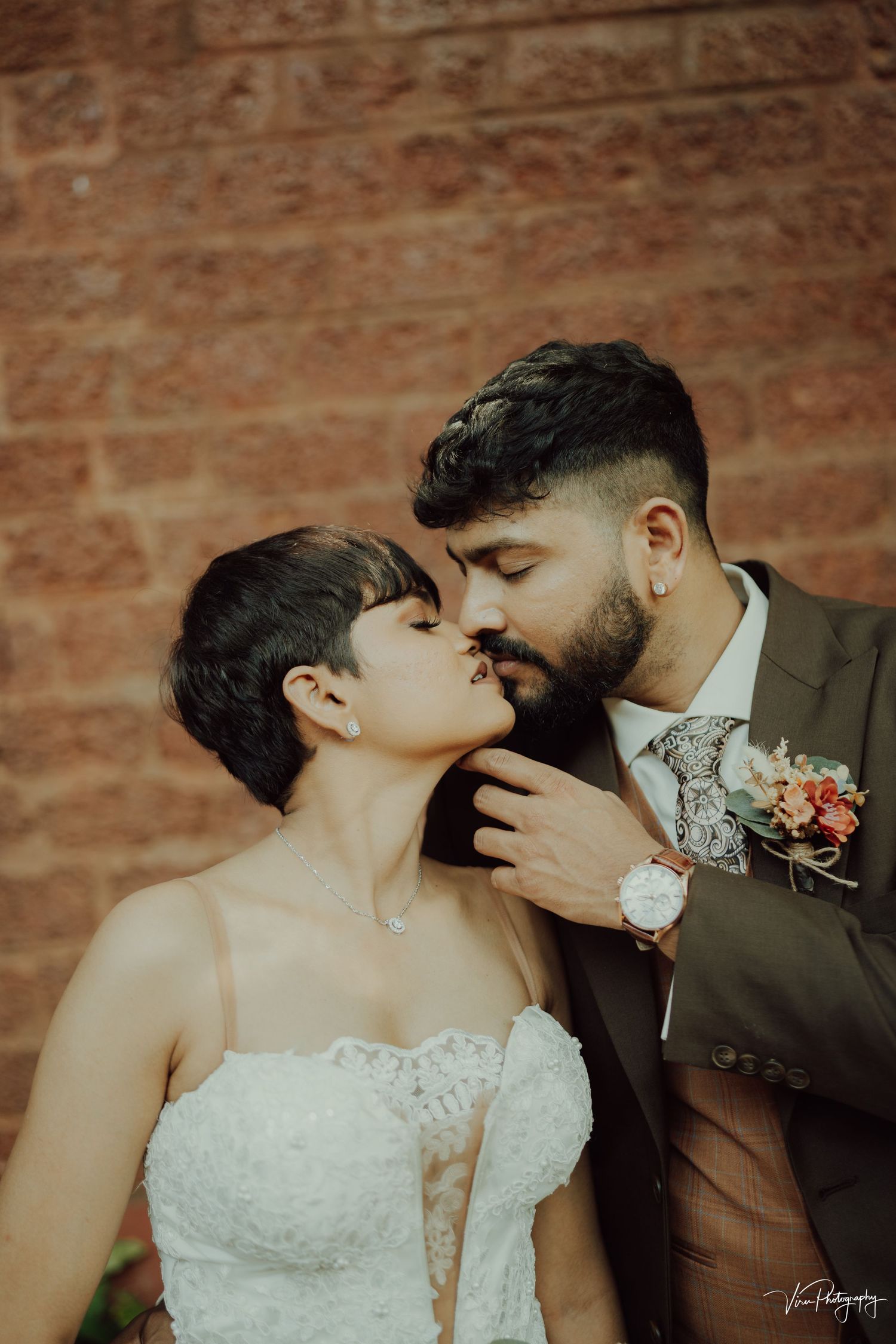 A romantic wedding couple sharing a tender kiss against a rustic brick wall backdrop with floral boutonniere details.