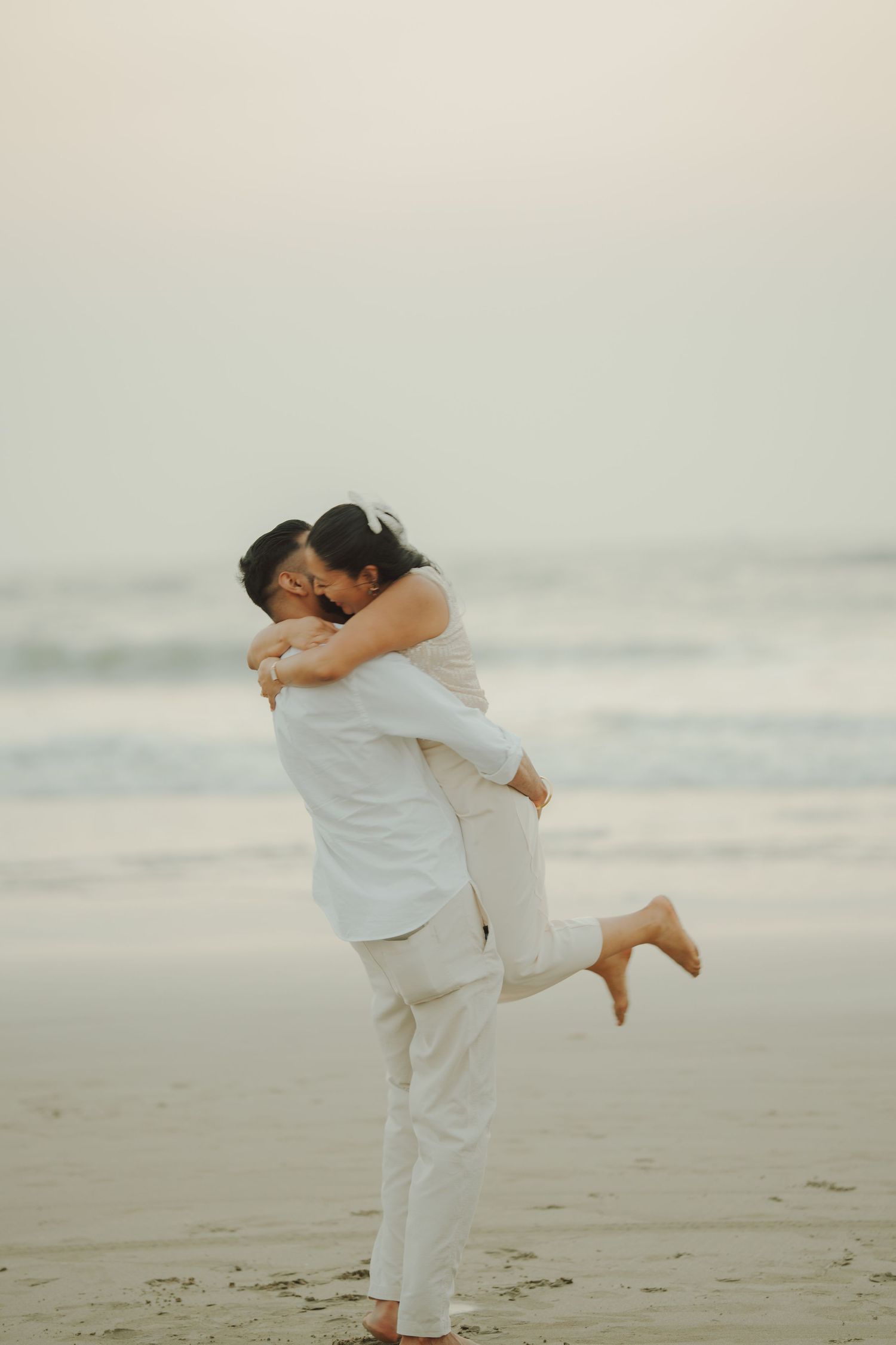 Couple in white outfits sharing a romantic kiss on a beach, man lifting woman as ocean waves roll in behind them.