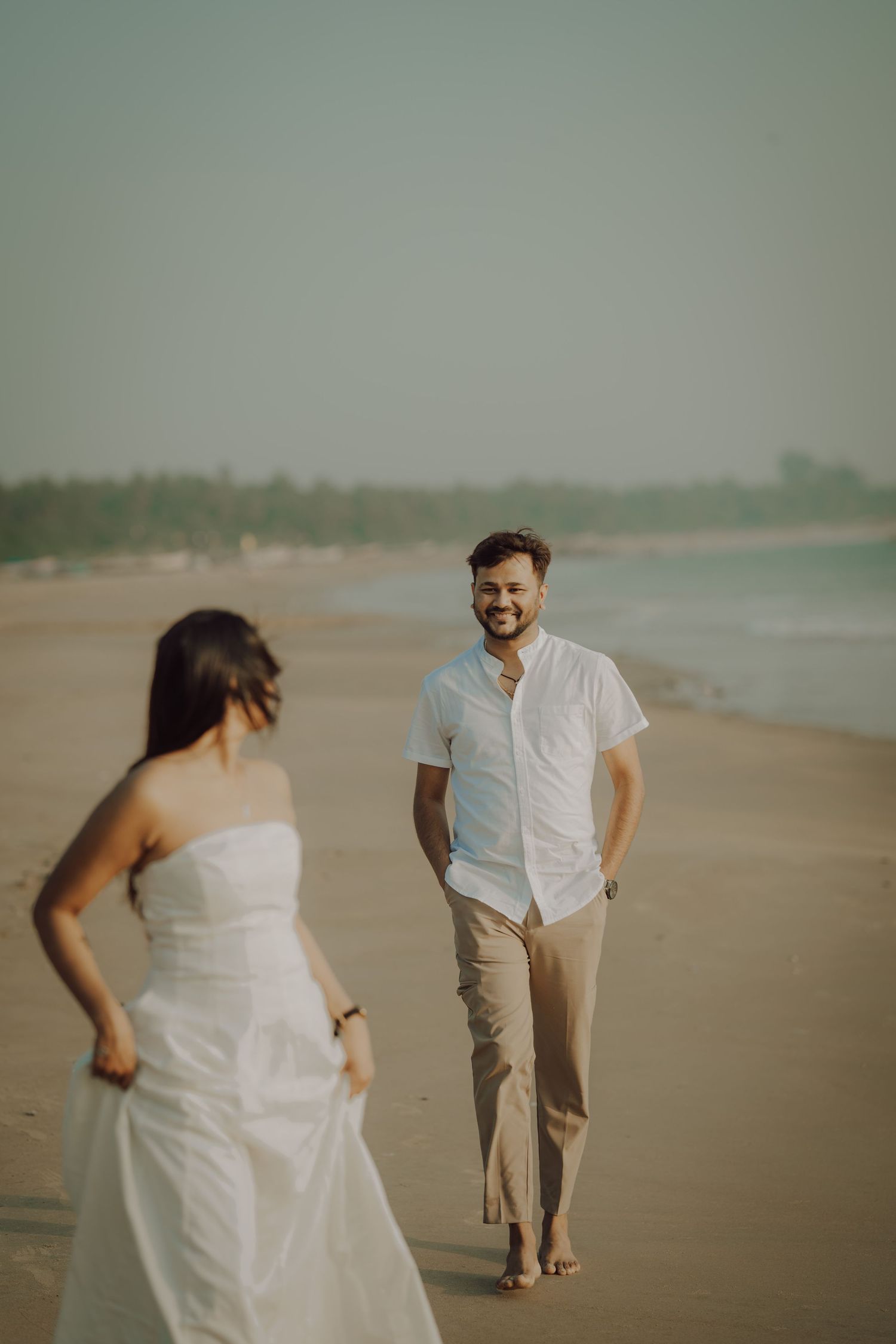 A couple in white attire walks playfully along a misty beach during a romantic photoshoot at sunset.