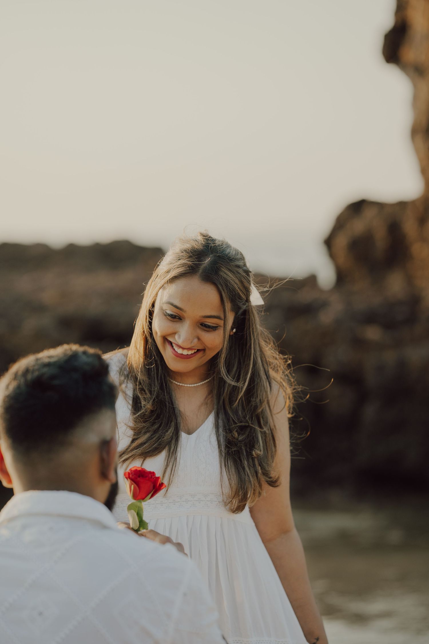 A romantic proposal scene at sunset with a red rose and white dress against a natural rocky backdrop.