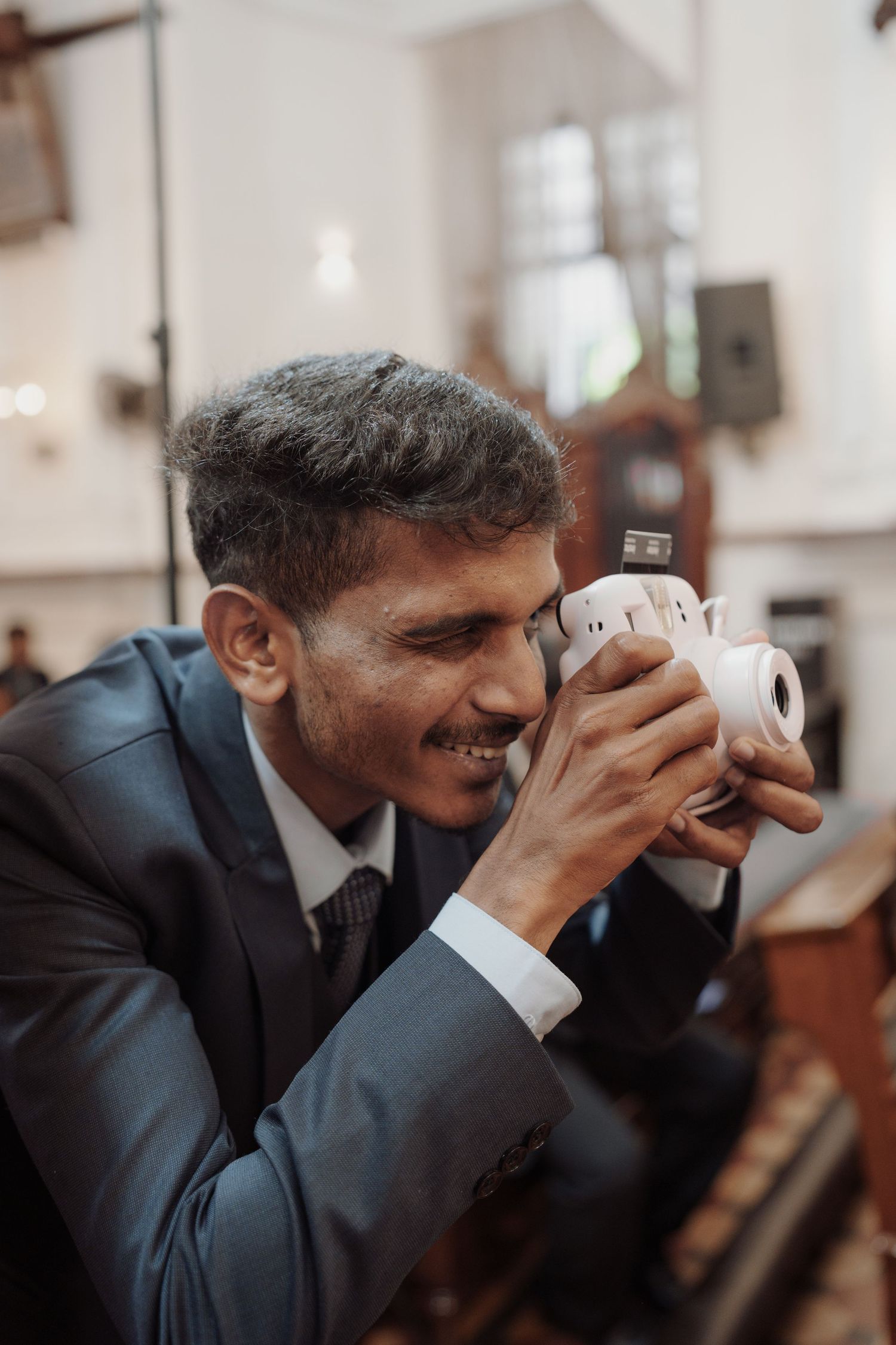 Smiling man in a gray suit takes a photo with a white instant camera inside a church.