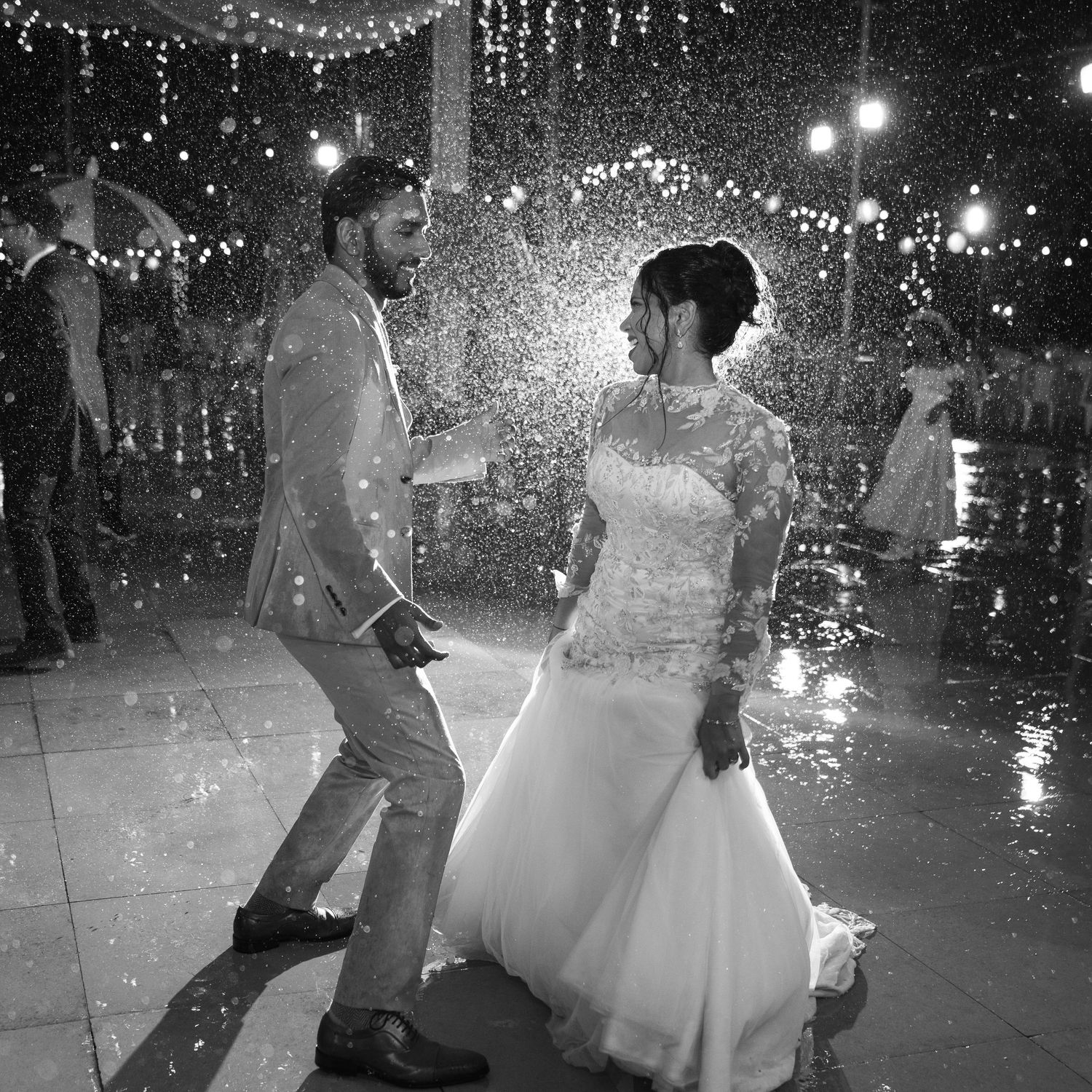 A black and white photo of a bride and groom dancing joyfully in a sparkling shower of water at their evening wedding reception.