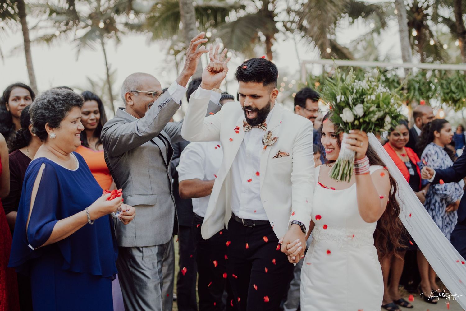 Wedding celebration outdoors with guests throwing confetti in celebration while palm trees sway in background.