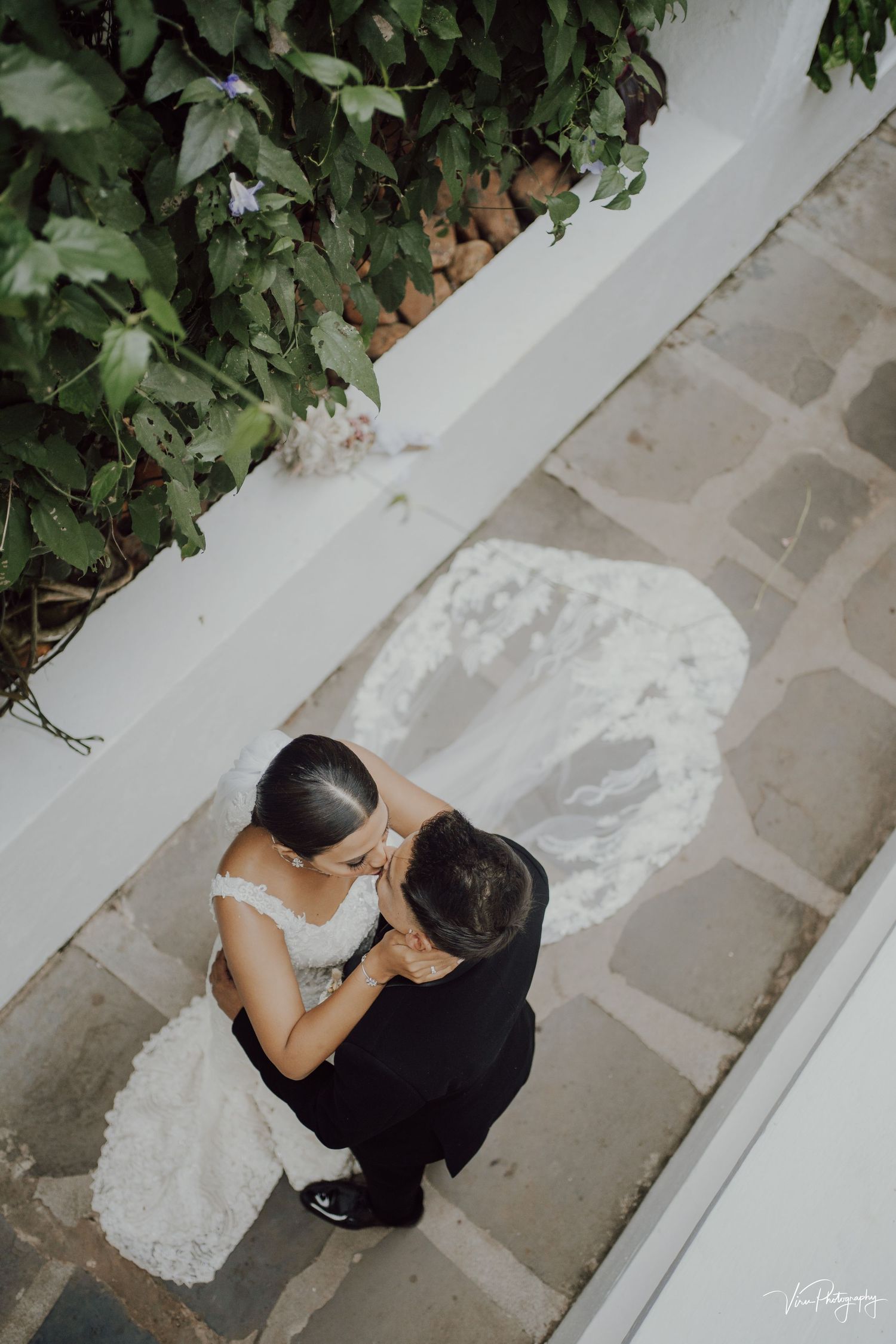 Aerial view of a couple in wedding attire embracing on a stone paved walkway beside green foliage and white walls.