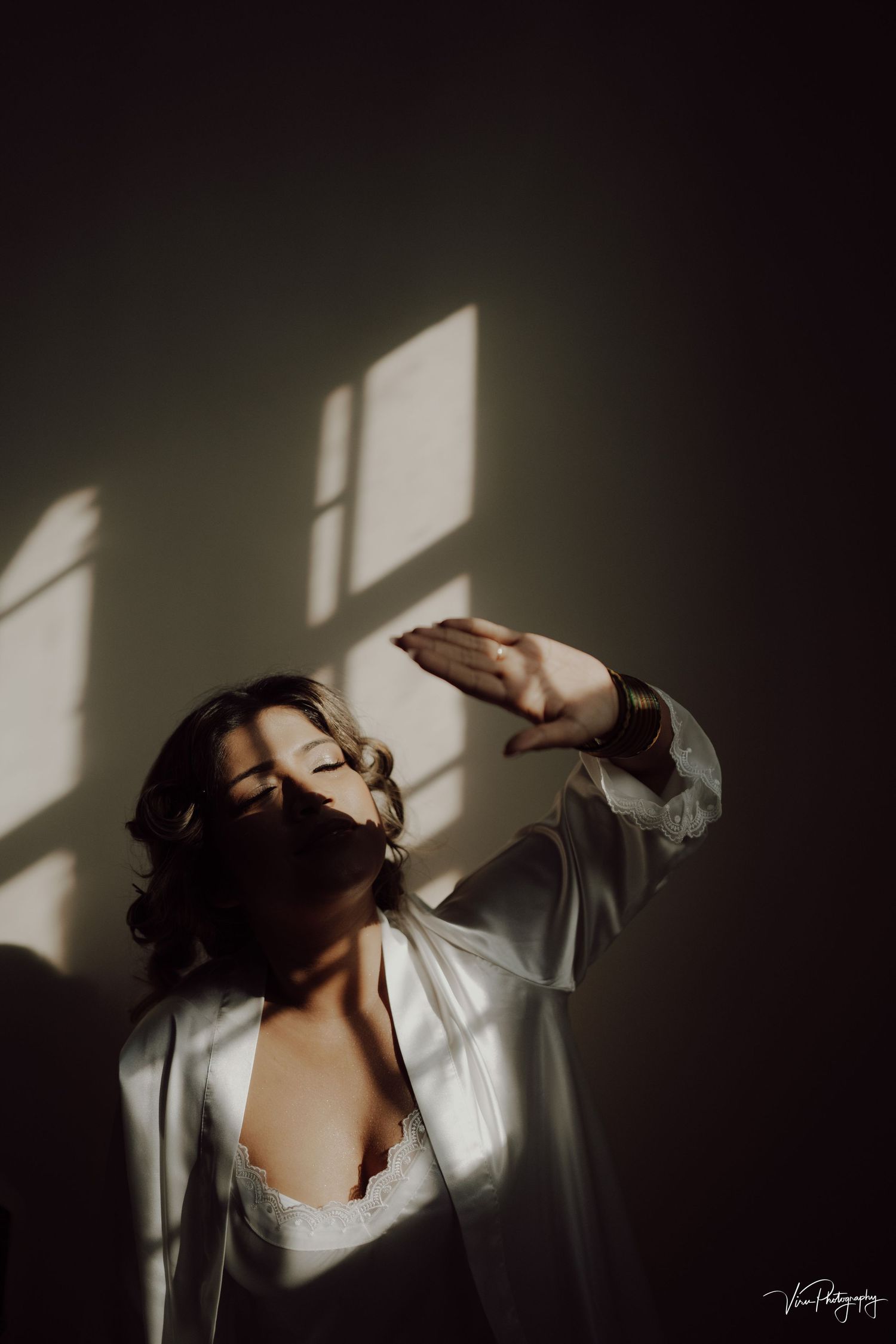 A moody portrait of a person in a white shirt with dramatic window light casting geometric shadows in a dark room.