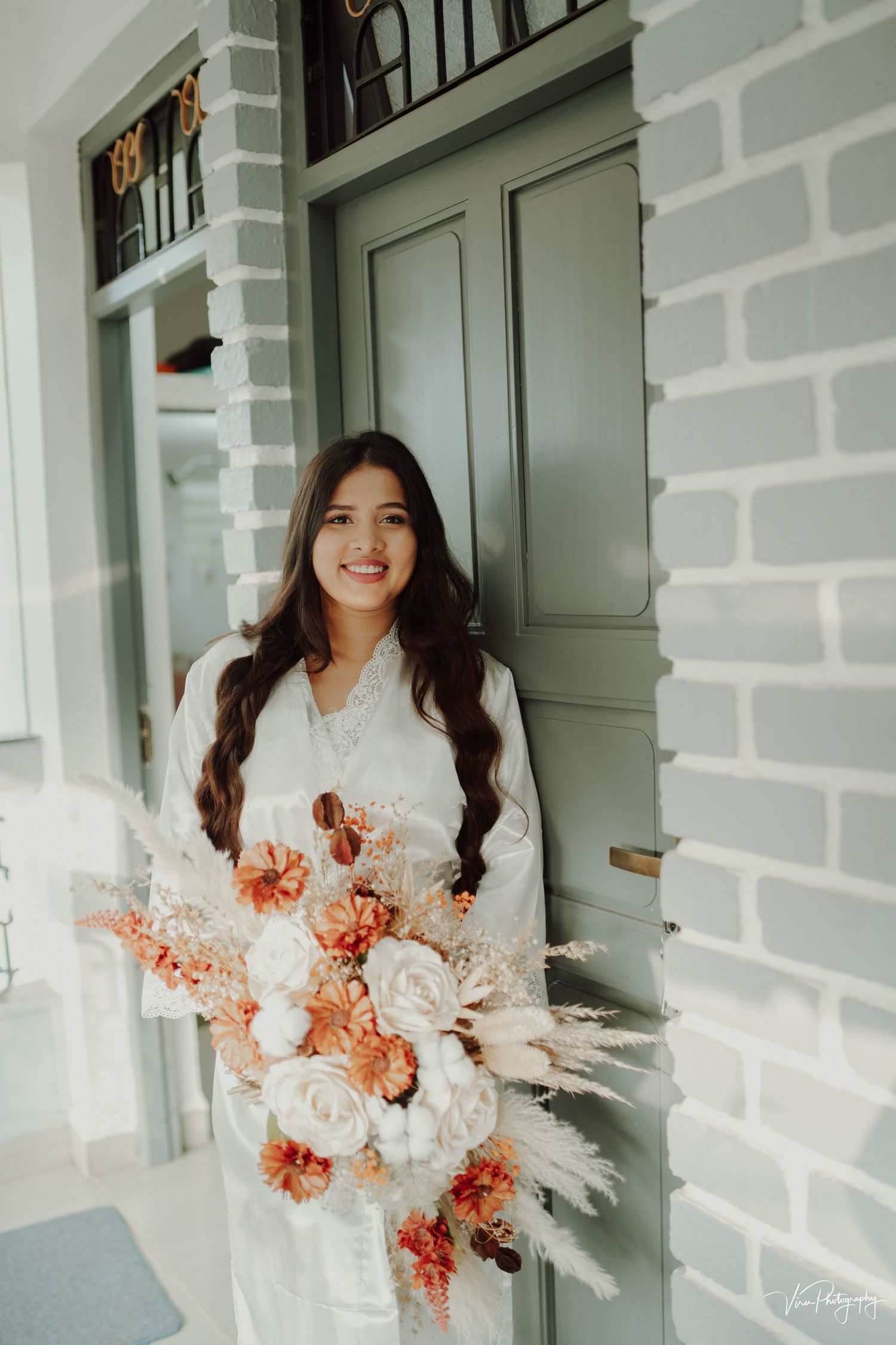 A person in a white dress holds a romantic bridal bouquet of peach and blush roses against a grey brick wall and green door.