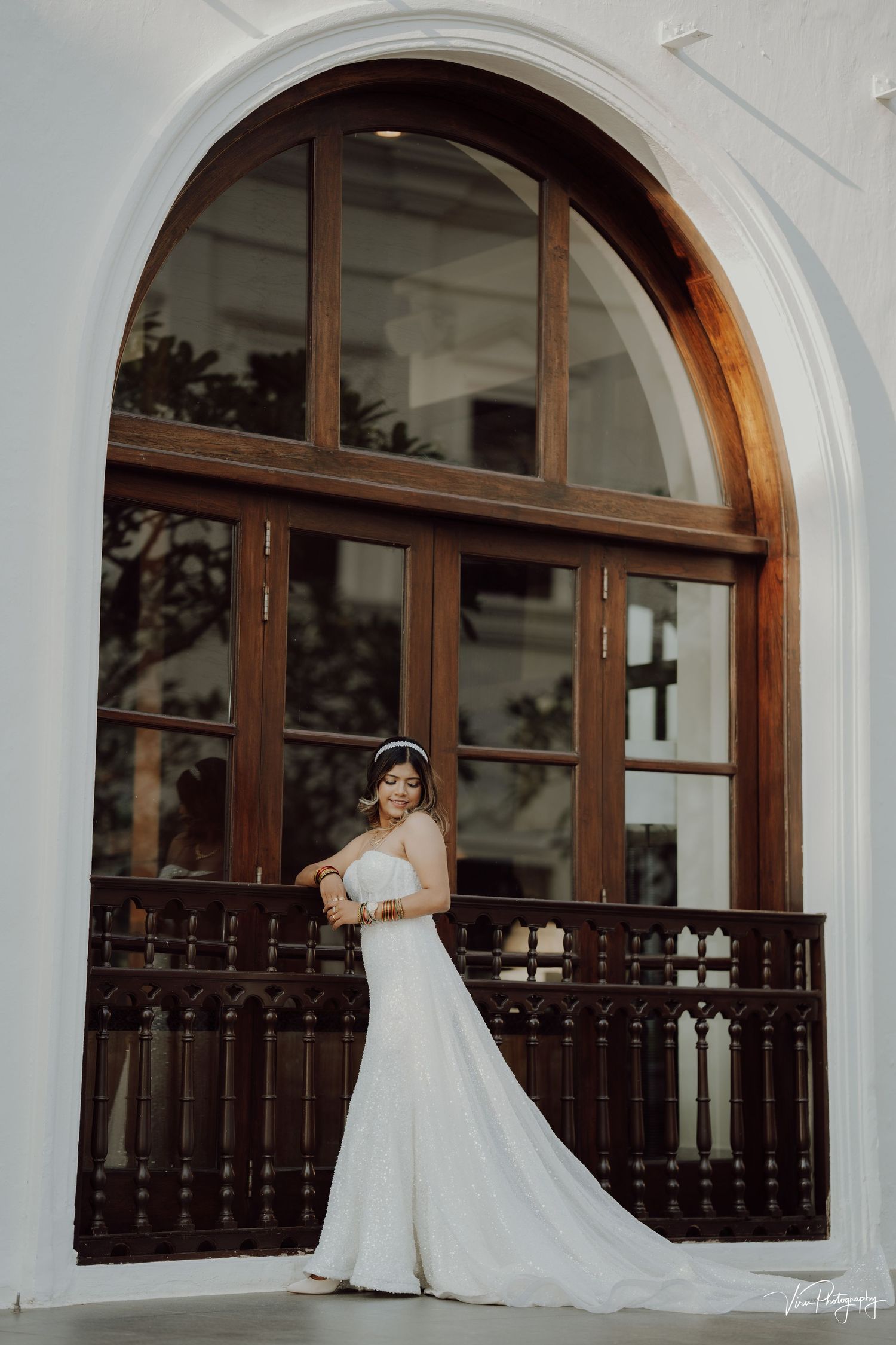 A bride in a flowing white wedding gown poses elegantly in front of a grand arched window with wooden details and railings.