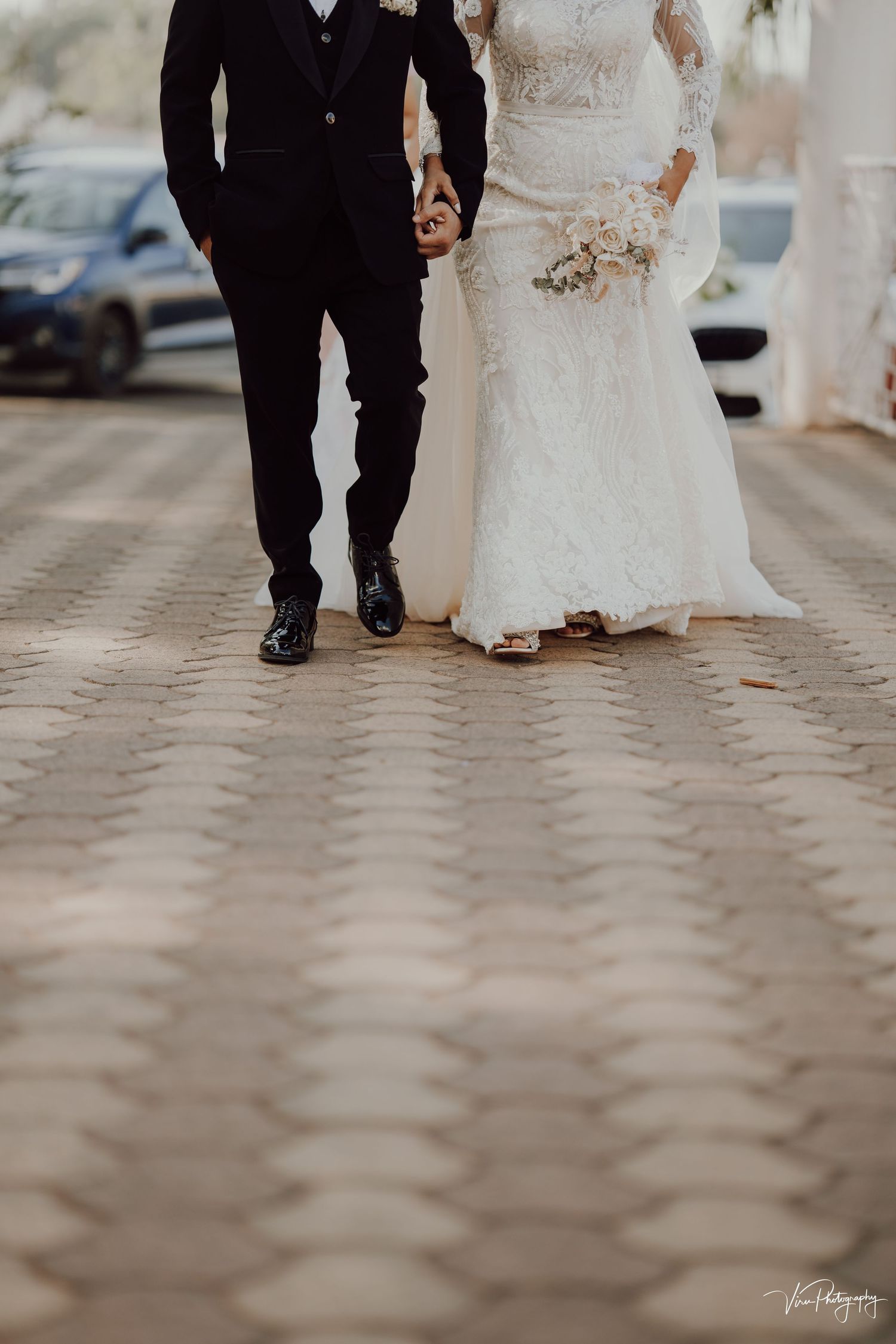 A romantic low-angle shot of a bride and groom walking together on a paved pathway, focusing on their legs and formal attire.