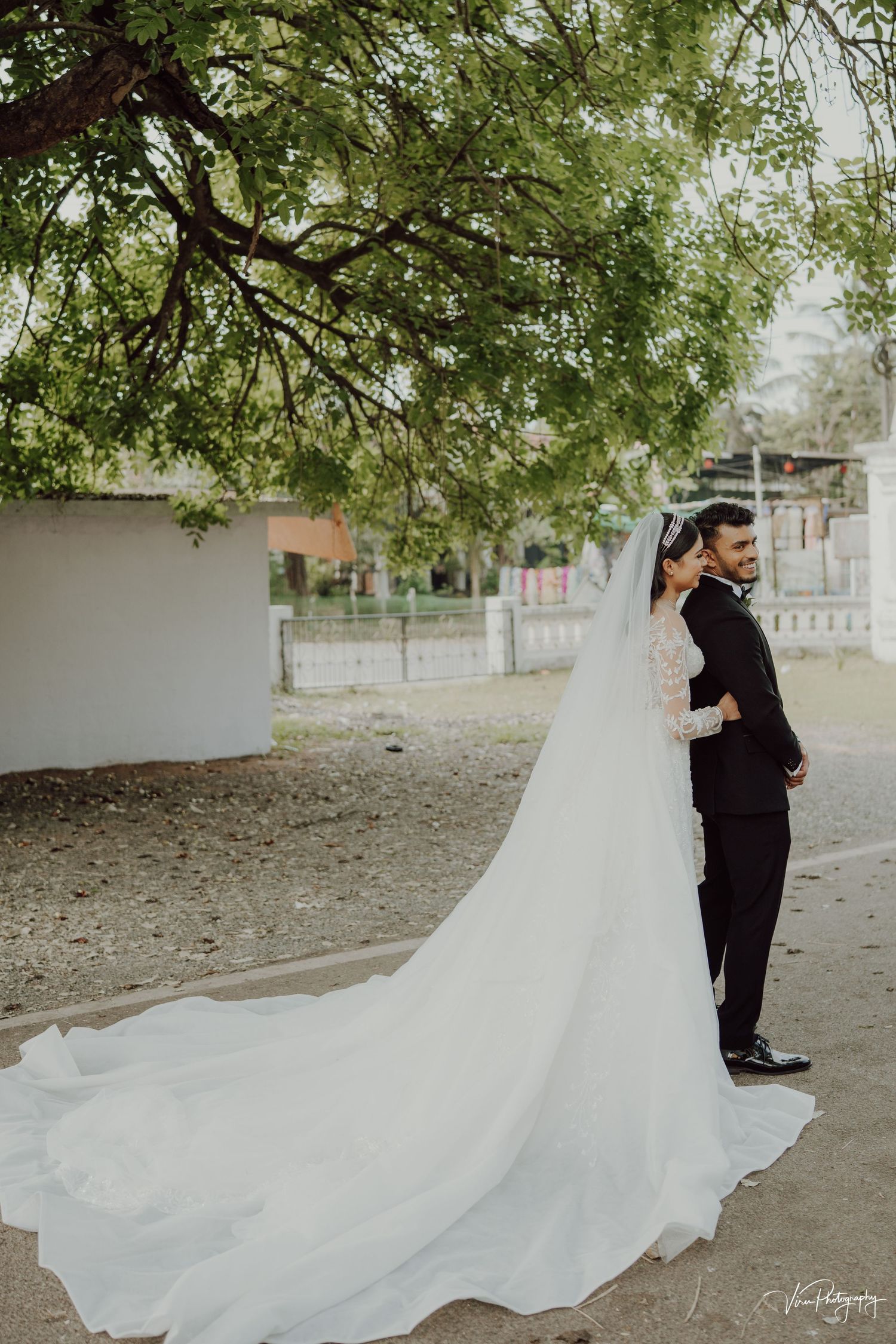Outdoor wedding photo showing a flowing white wedding gown with a dramatic long train under a tree canopy.