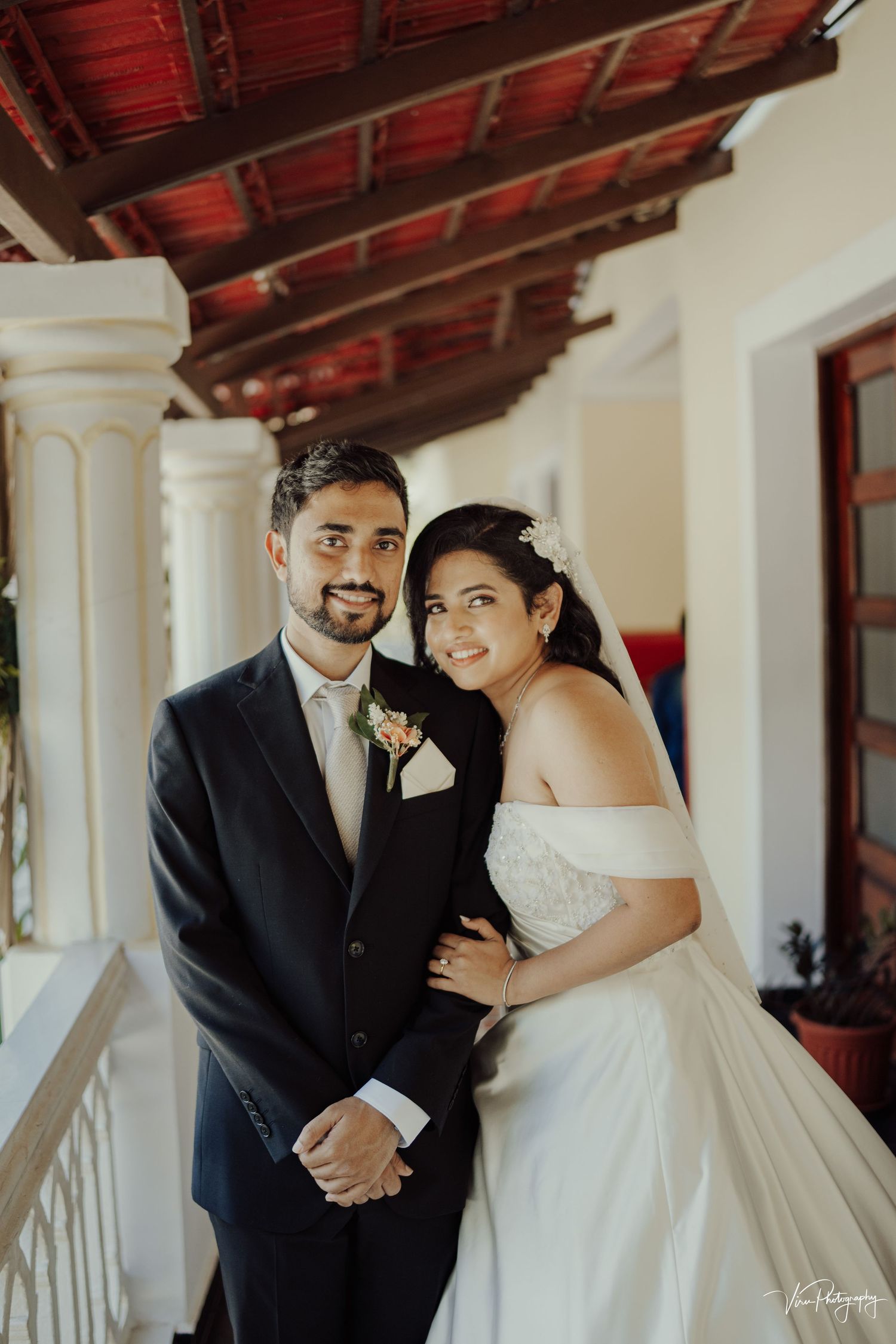 A couple poses together on their wedding day in front of white columns and wooden beams at a venue.