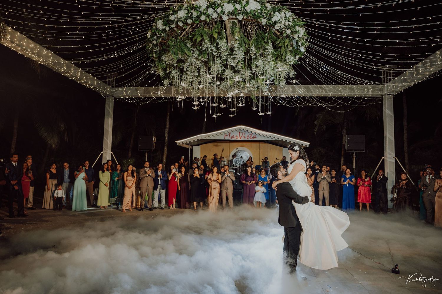 Couple shares first dance surrounded by wedding guests under twinkling lights and fog at an outdoor reception.