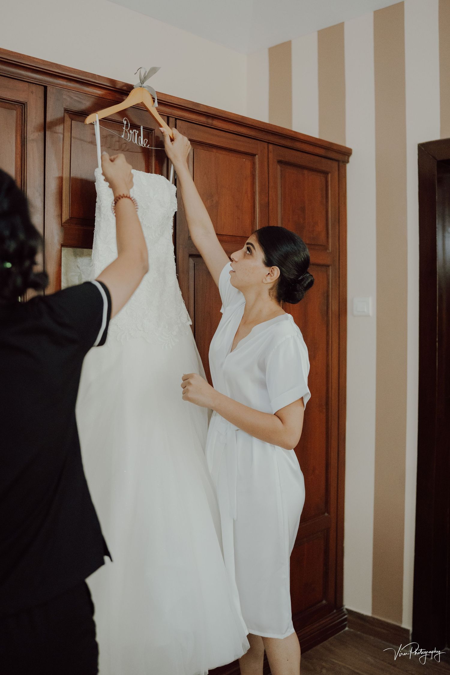 Two people hang up an elegant white wedding dress on a wooden hanger against a wall with striped wallpaper.