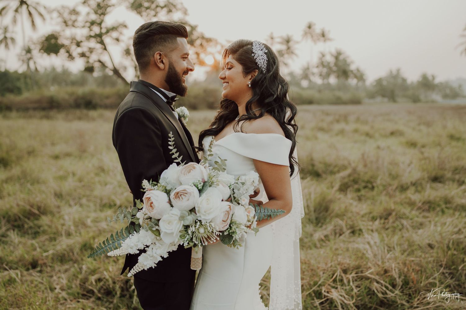 A romantic wedding photo in a grassy field at sunset with palms in the background and a white floral bouquet.