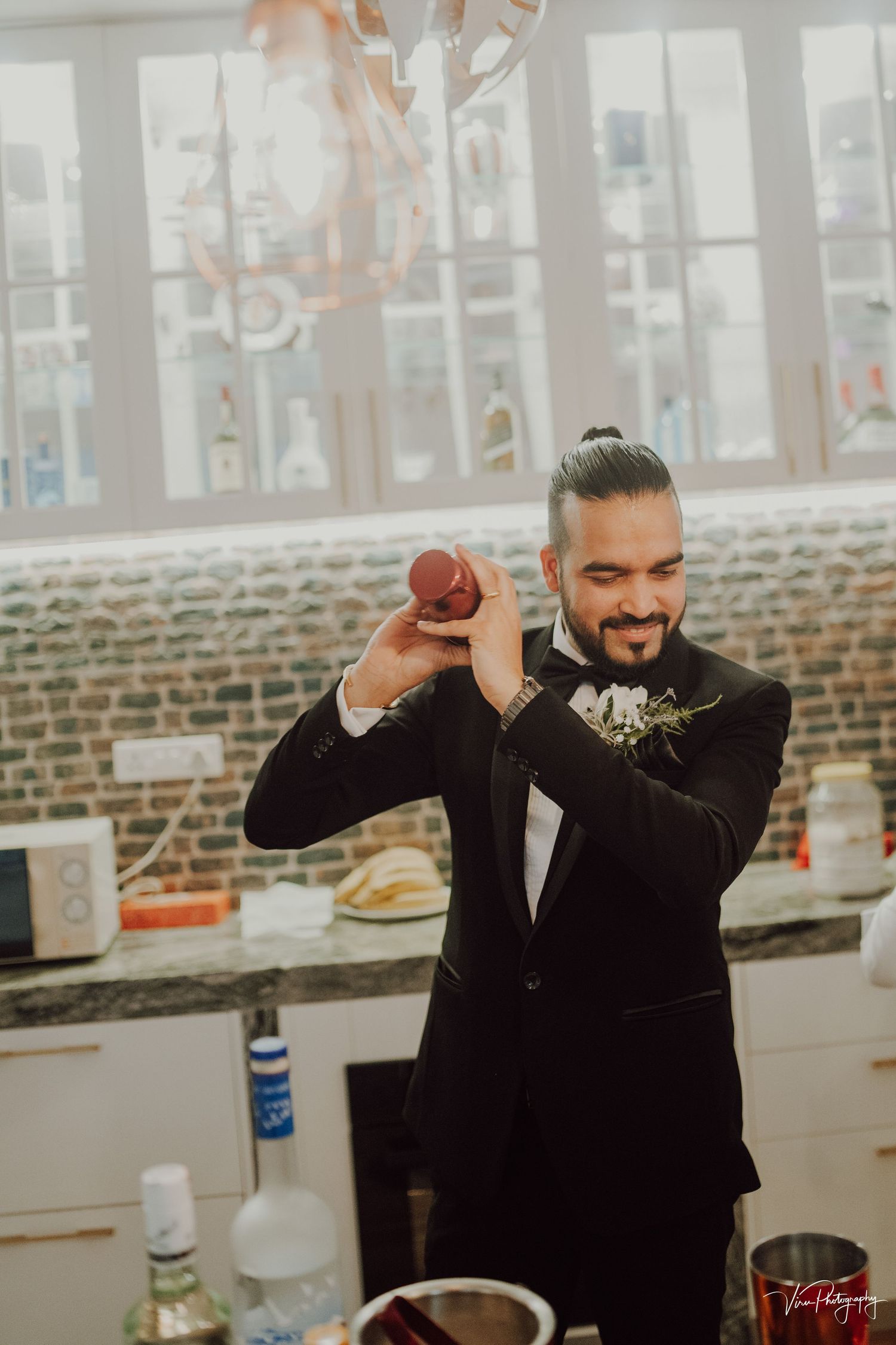 Groom in black tuxedo and boutonniere playfully shakes a cocktail shaker at an indoor reception bar.