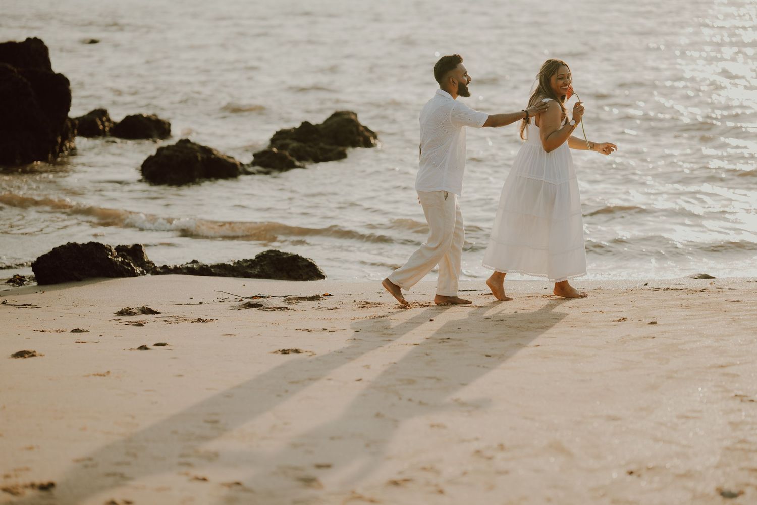 Two people walk hand in hand along a scenic beach with rocky outcrops and gentle waves at sunset.