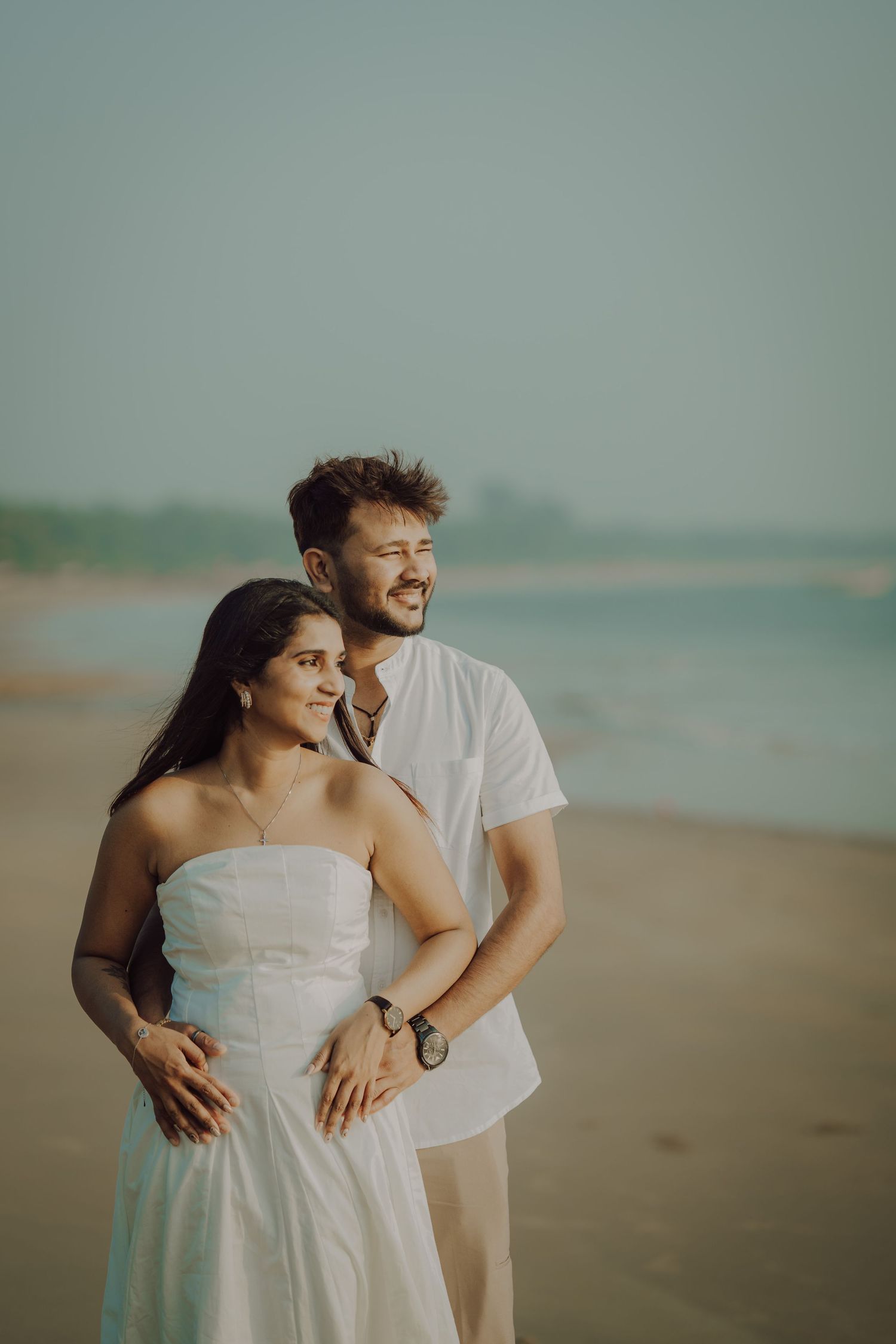 A romantic couple embraces and laughs together while walking along a misty beach in white attire.