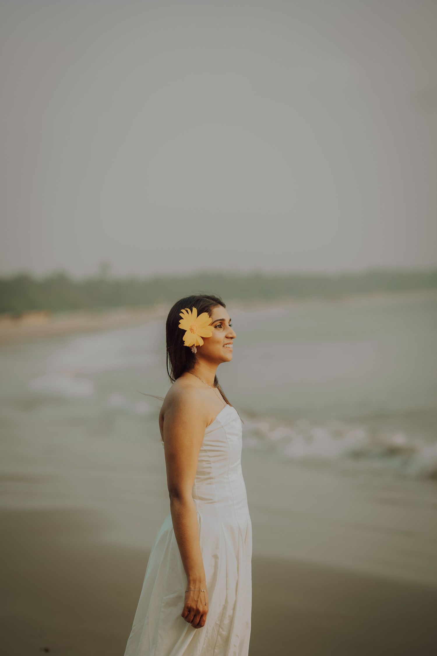 A person in a white dress stands contemplatively on a misty beach during an overcast day.