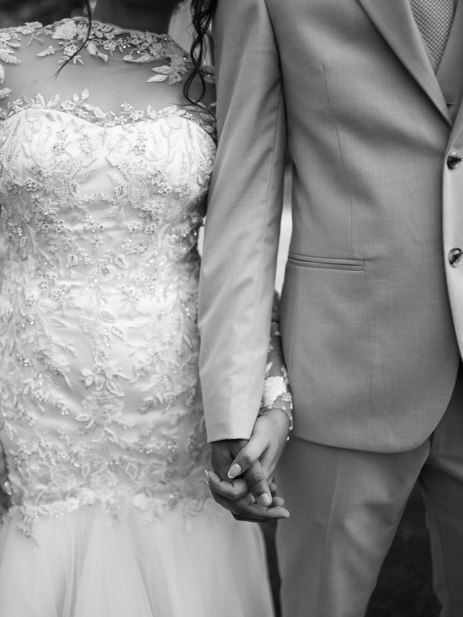 Black and white close-up of a bride in a lace gown holding hands with a groom in a suit.