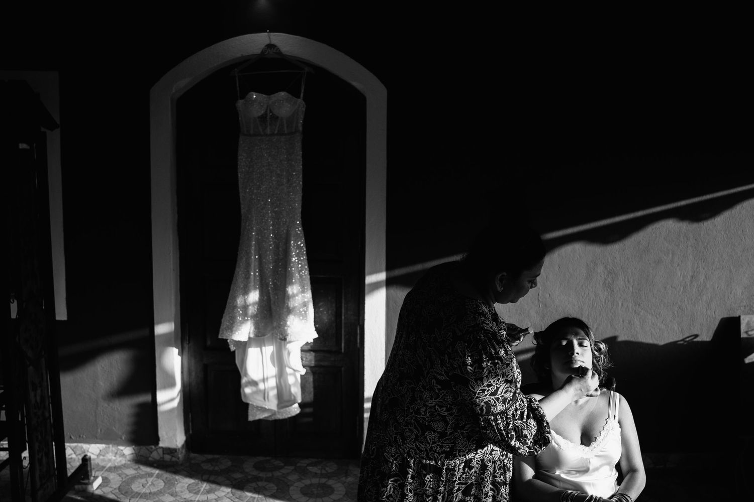 A moody black and white photo showing a dress hanging in a doorway while someone sits nearby in shadow.
