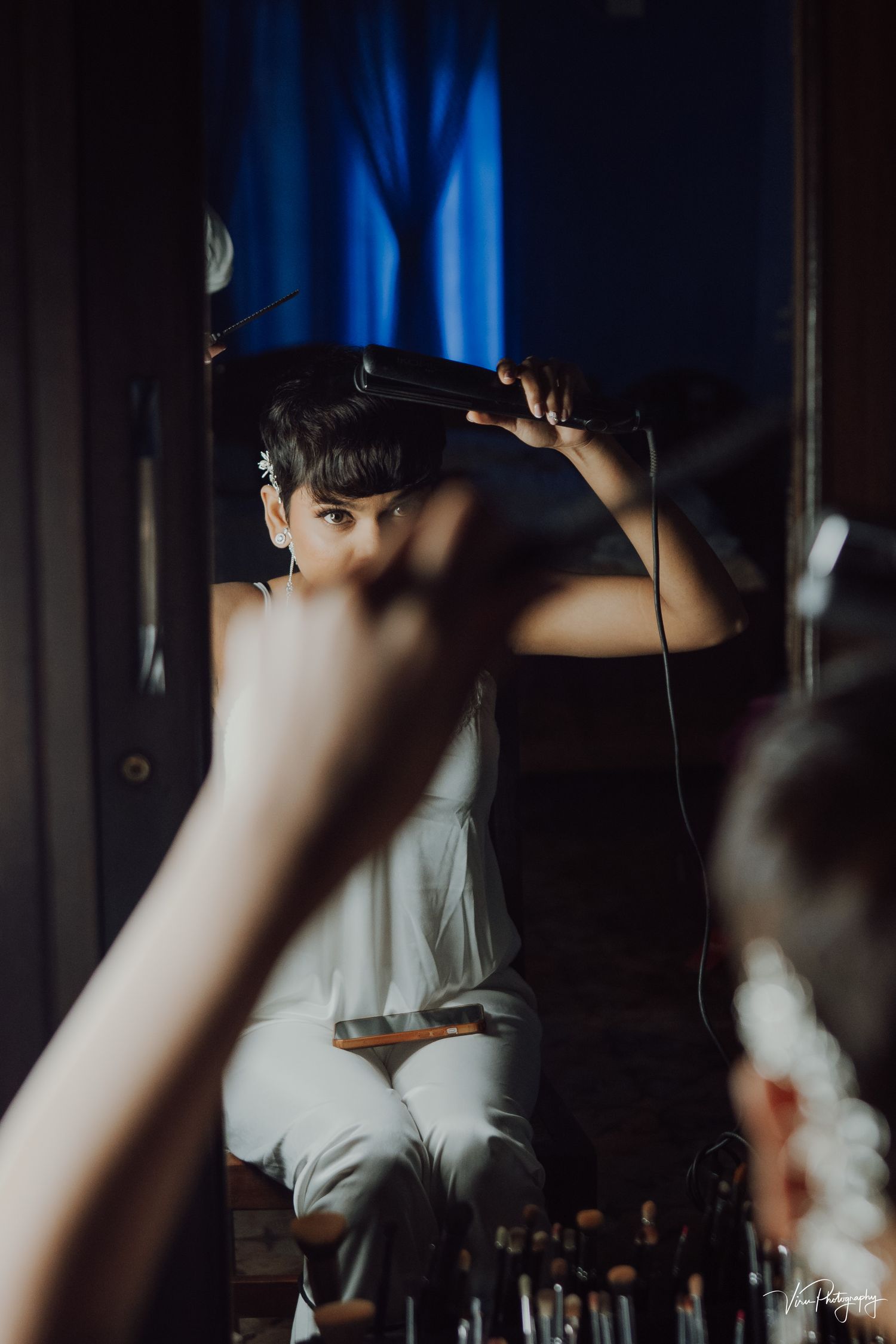 Behind-the-scenes shot of a person in white getting hair styled with dramatic blue lighting in a makeup room.