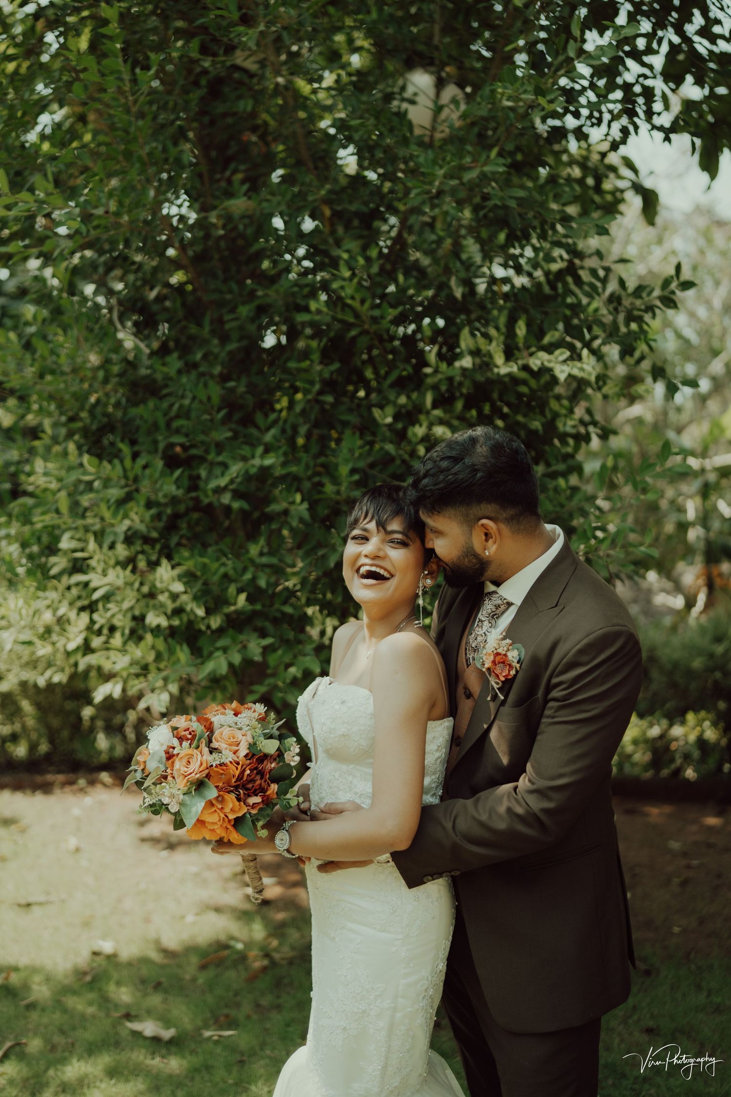 A joyful wedding couple shares a candid moment together outdoors with orange floral bouquet and natural greenery backdrop.
