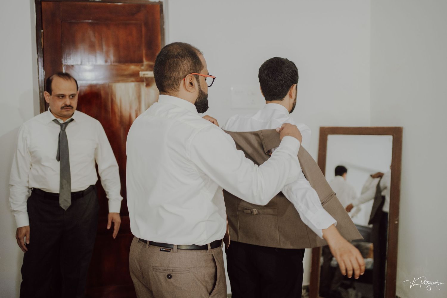 Three people in formal attire getting ready in a light-filled room with a wooden door and mirror visible.