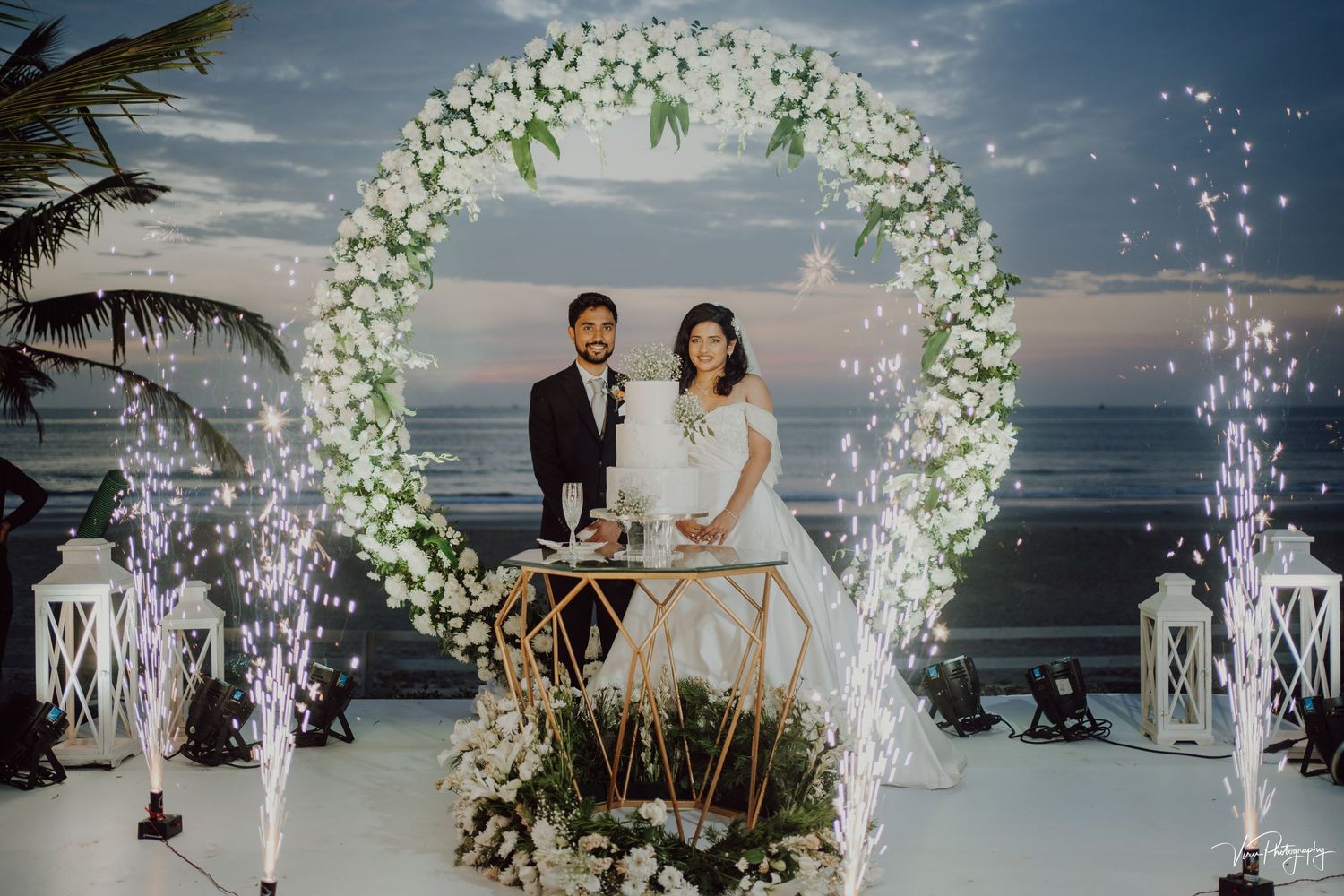 A romantic beach wedding ceremony setup with a circular floral arch and firework sparklers against an ocean sunset backdrop.