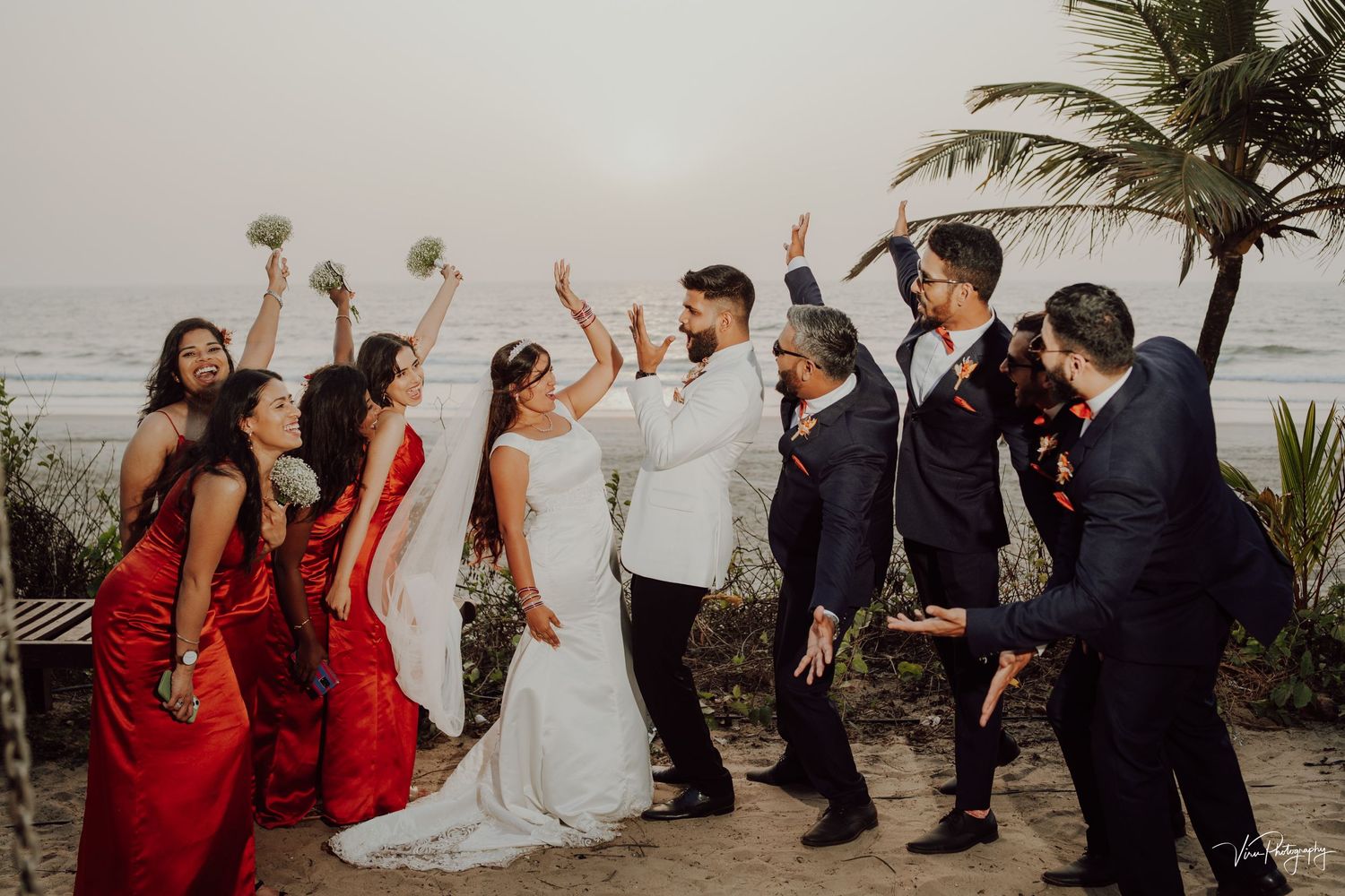 Wedding party celebrates on a beach with palm trees as guests wave their hands in celebration at a sunset ceremony.