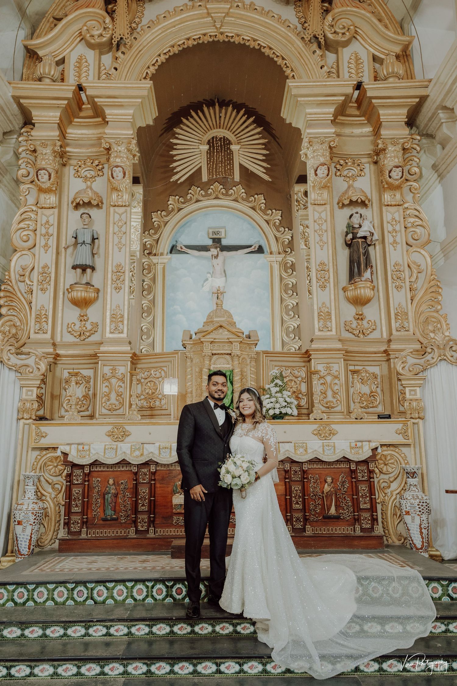 A couple stands in front of an ornate baroque church altar with gilded decorations and religious sculptures.