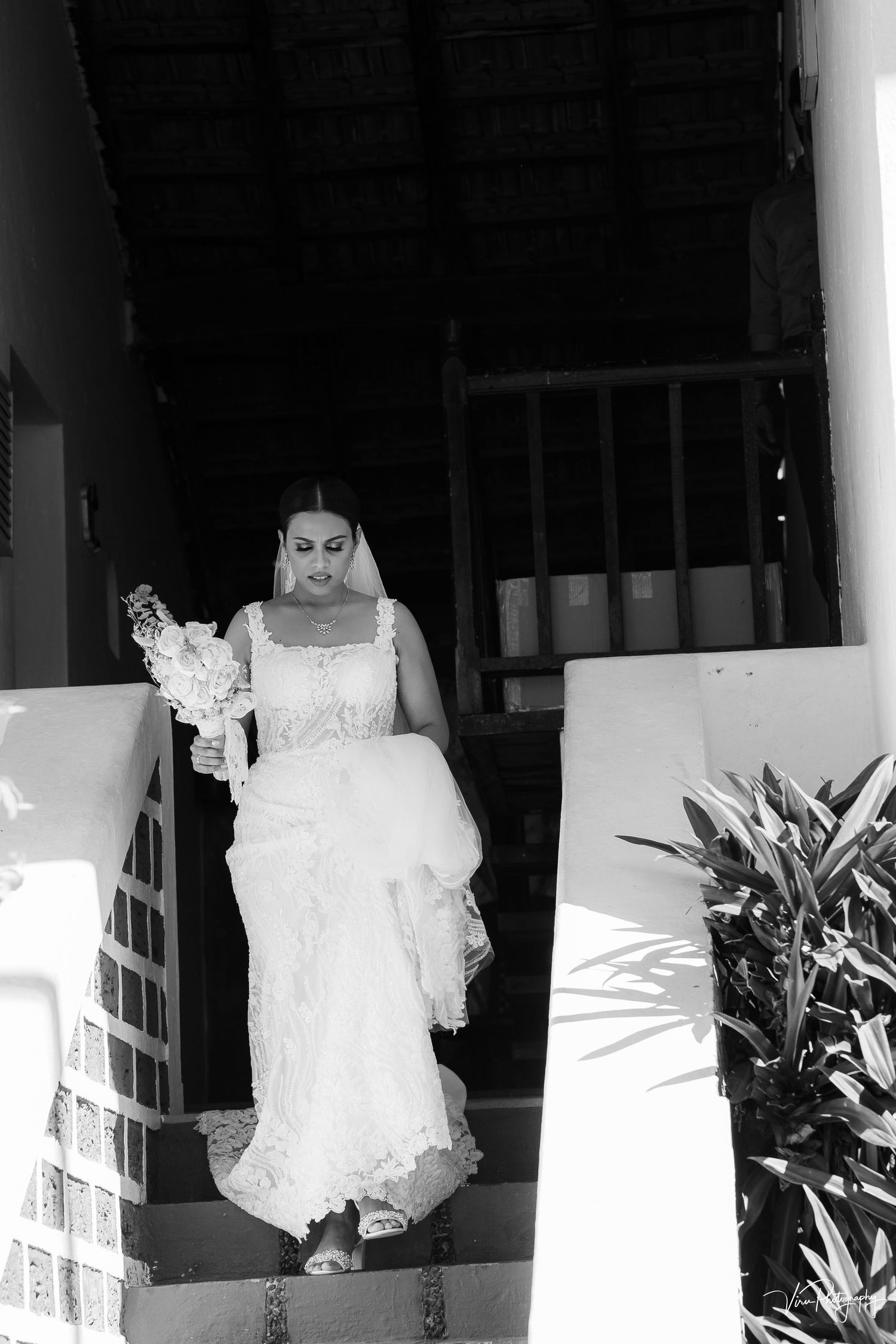 A bride in an elegant white wedding dress descends stairs at a botanical venue in black and white photography.