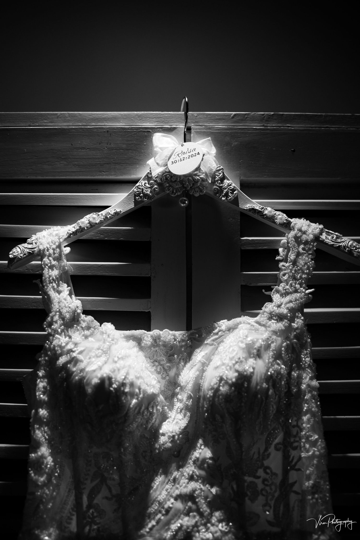 A black and white close-up of a lace wedding dress hanging on a wooden hanger in dramatic lighting.