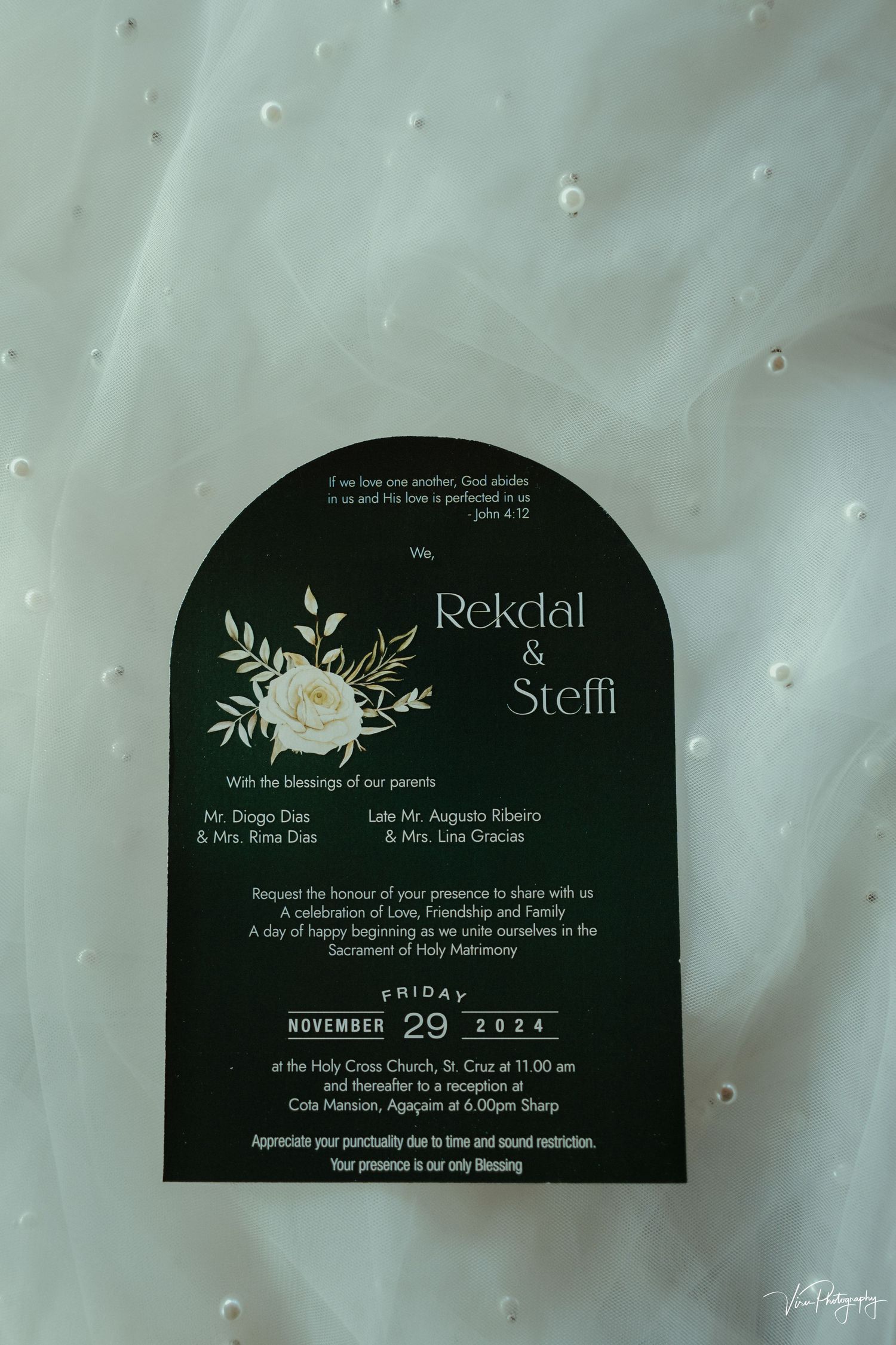 A black arched wedding invitation with silver text and white floral design rests on a textured white surface.