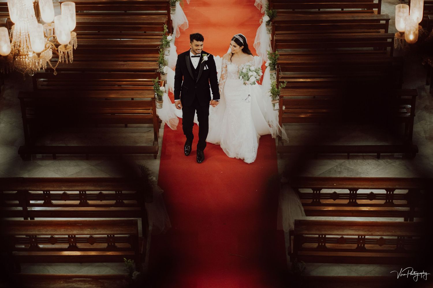 Romantic overhead view of a wedding ceremony in a church with red carpet and wooden pews illuminated by chandeliers.